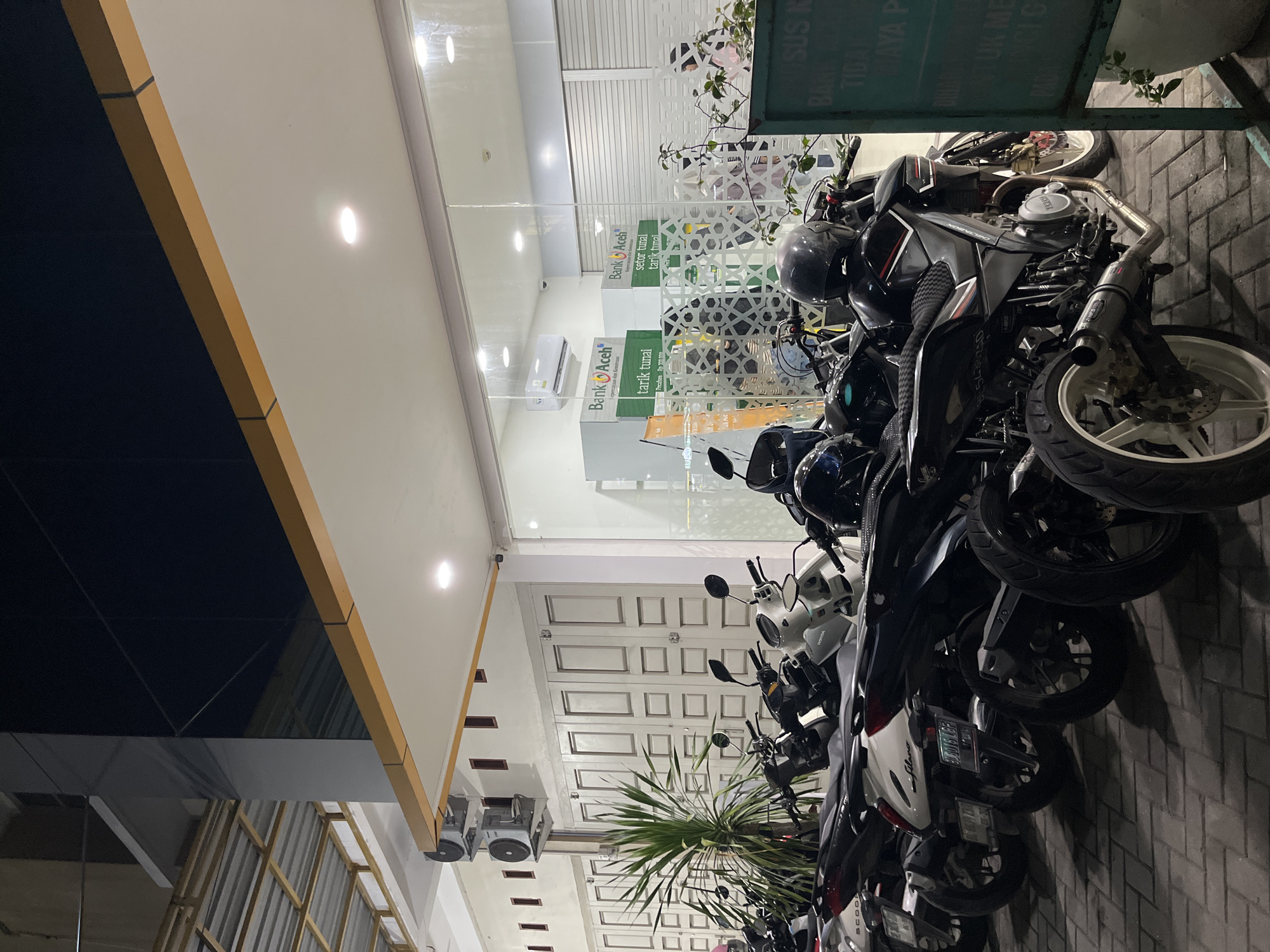A nighttime, eye-level medium shot shows a row of parked motorcycles in front of a building with a glass facade. The building appears to be a Bank Aceh branch, as indicated by signage visible through the glass. Several motorcycles are parked in the foreground, with their headlights and taillights off. The motorcycles are of various makes and models, with some being scooters and others sport bikes. Behind the motorcycles, the building features a large glass window that offers a view into the bank's interior. Inside the bank, ATMs are visible, as well as what appear to be closed teller windows. The building has a modern architectural style, with clean lines and a prominent yellow awning extending over the entrance. The lighting in the scene is dim, with the primary light sources being the interior lights of the bank and some overhead lighting from the building's exterior. The ground is paved with interlocking bricks. A tall, leafy green plant is positioned to the left of the frame.