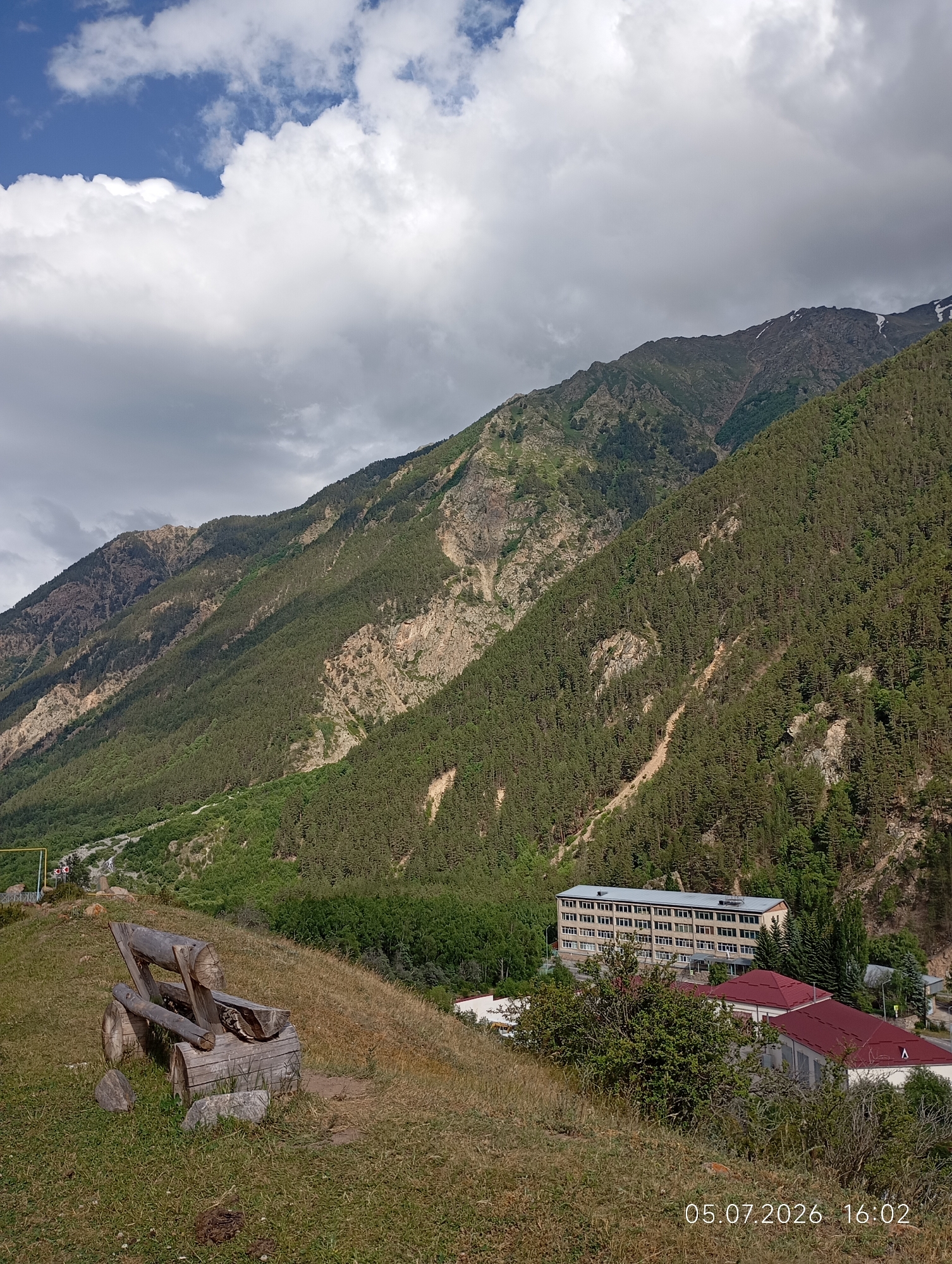 A wide-angle, high-angle outdoor photograph depicts a mountain landscape with a building complex visible in the mid-ground. In the foreground, a rustic wooden bench made of logs sits on a grassy slope, accompanied by a few scattered rocks. The bench is empty. Behind the bench, the terrain slopes down towards a valley. The mid-ground features a large, multi-story building with a light-colored facade and many windows, surrounded by trees. Adjacent to this building is another structure with a red roof. The background is dominated by steep, densely forested mountains under a sky with scattered clouds. The date "05.07.2026 16:02" is superimposed in the lower right corner of the image. This location is identified as the city of Neutrino, Russia.