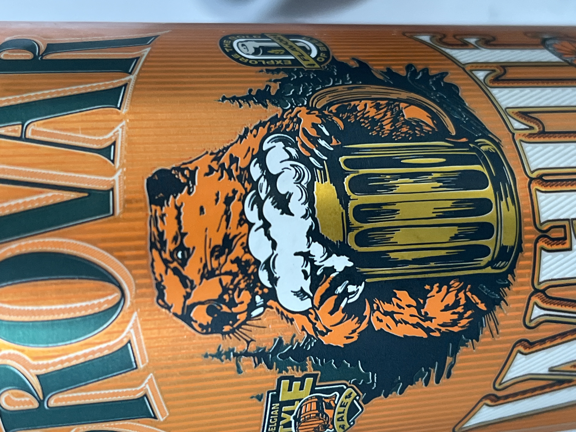 The image shows a close-up of a can of beer with an orange background and vertical lines. A stylized orange beaver with a black outline is depicted holding a frothy mug of beer. To the left of the beaver, there is a rectangular badge with green and yellow accents that says "BELGIAN STYLE ALE". To the right of the beaver, there is a circular badge with white and yellow accents that says "EXPLORE THE FLAVOR OF BEER". The word "BAR" is visible in large white letters at the bottom of the image. The image does not depict any specific location in the city of Il'ino, Russia.