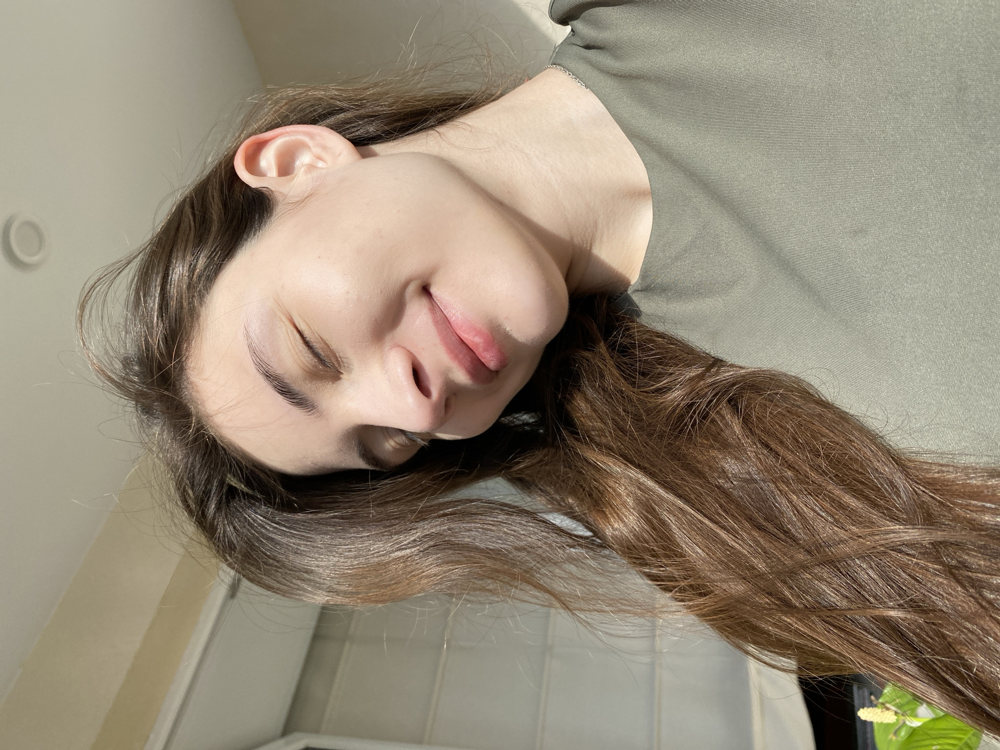The image captures a young woman, likely in Votkinsk, Russia, illuminated by bright natural sunlight indoors. She is positioned as if lying down or reclining, viewed from a slightly elevated angle. Her eyes are closed, and a gentle smile plays on her lips, conveying an expression of peace, relaxation, or contentment. She has long, wavy, medium-brown hair with lighter, sun-kissed highlights, which cascades around her head. She is wearing a simple, olive-green top with a fine, ribbed texture, and a faint silver chain is visible on her neck.
Strong, direct sunlight highlights her features and hair, casting distinct shadows on her neck and under her chin, indicating a light source, likely a window, to her side. The background consists primarily of light-colored walls. In the upper left, a faint circular object is visible on the wall, possibly a light fixture or a reflective element. To the bottom right, a green plant with small white flowers adds a touch of nature to the indoor setting. The bright illumination strongly suggests it is daytime with sunny weather outside. There is no visible text in the image.