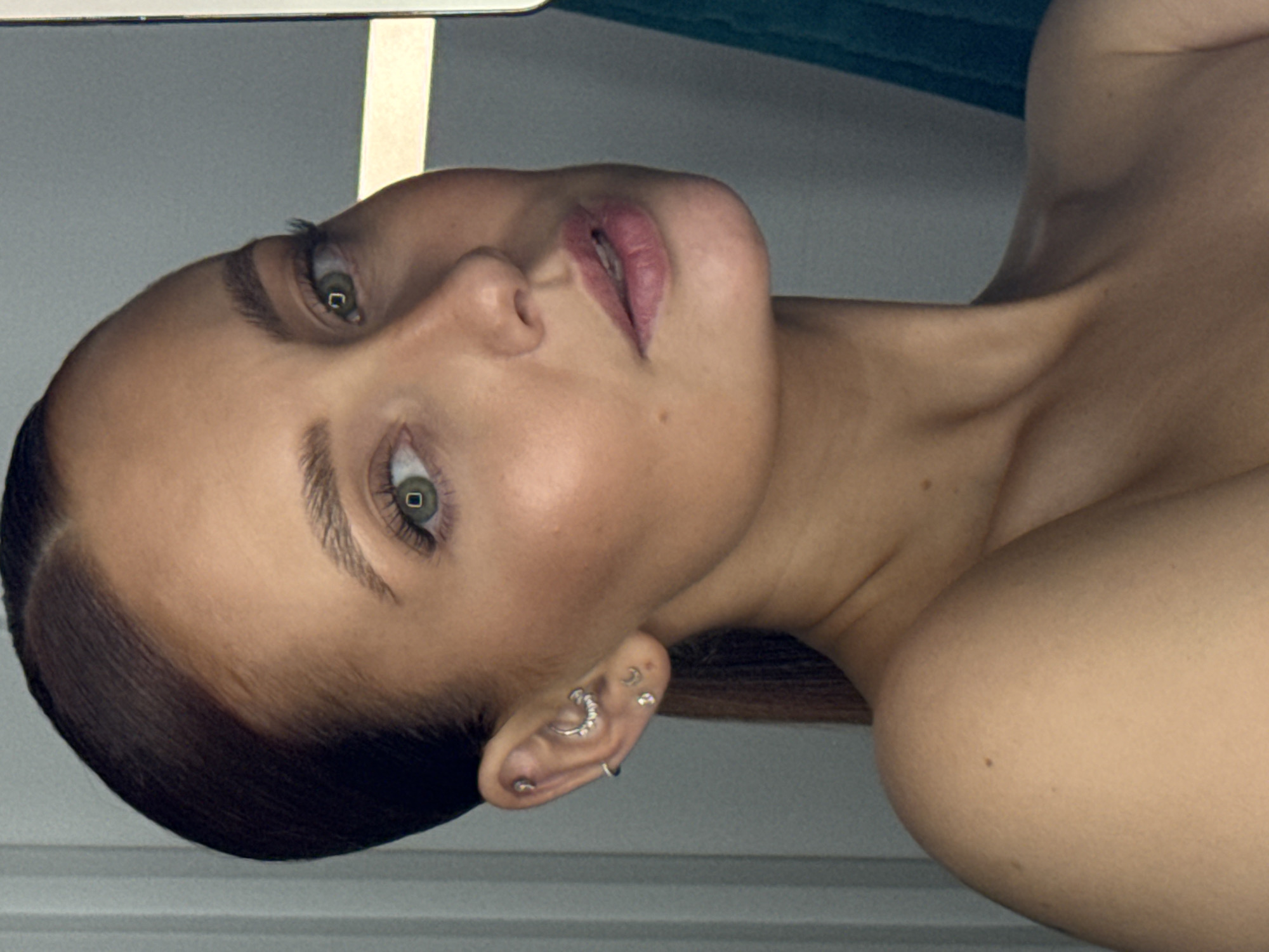 The media file displays a close-up portrait of a person, likely a young woman, with her head slightly tilted and her gaze directly engaging the viewer. She has fair skin, dark hair pulled back from her face, and striking emerald green eyes with visible highlights in her pupils, suggesting artificial light. Her lips are full and appear to have a light, natural-toned lipstick, slightly parted to reveal a glimpse of teeth.
The visible ear is adorned with multiple intricate piercings, including a crescent-shaped earring studded with stones along the helix, several small studs, and a simple hoop on the lobe. The subject's bare shoulders and neck are visible, showing smooth skin and the subtle definition of her collarbone.
The activity captured is a posed portrait. The setting appears to be indoors, against a minimalist and softly blurred background, consisting primarily of muted grey tones with a darker, possibly teal or green, surface visible in the upper right. The lighting is controlled, highlighting the subject's features. There are no specific location cues visible within the image itself to indicate Chaikovsky, Russia, nor is the time of day or weather discernible. No visible text is present. The overall mood conveyed is calm and direct.