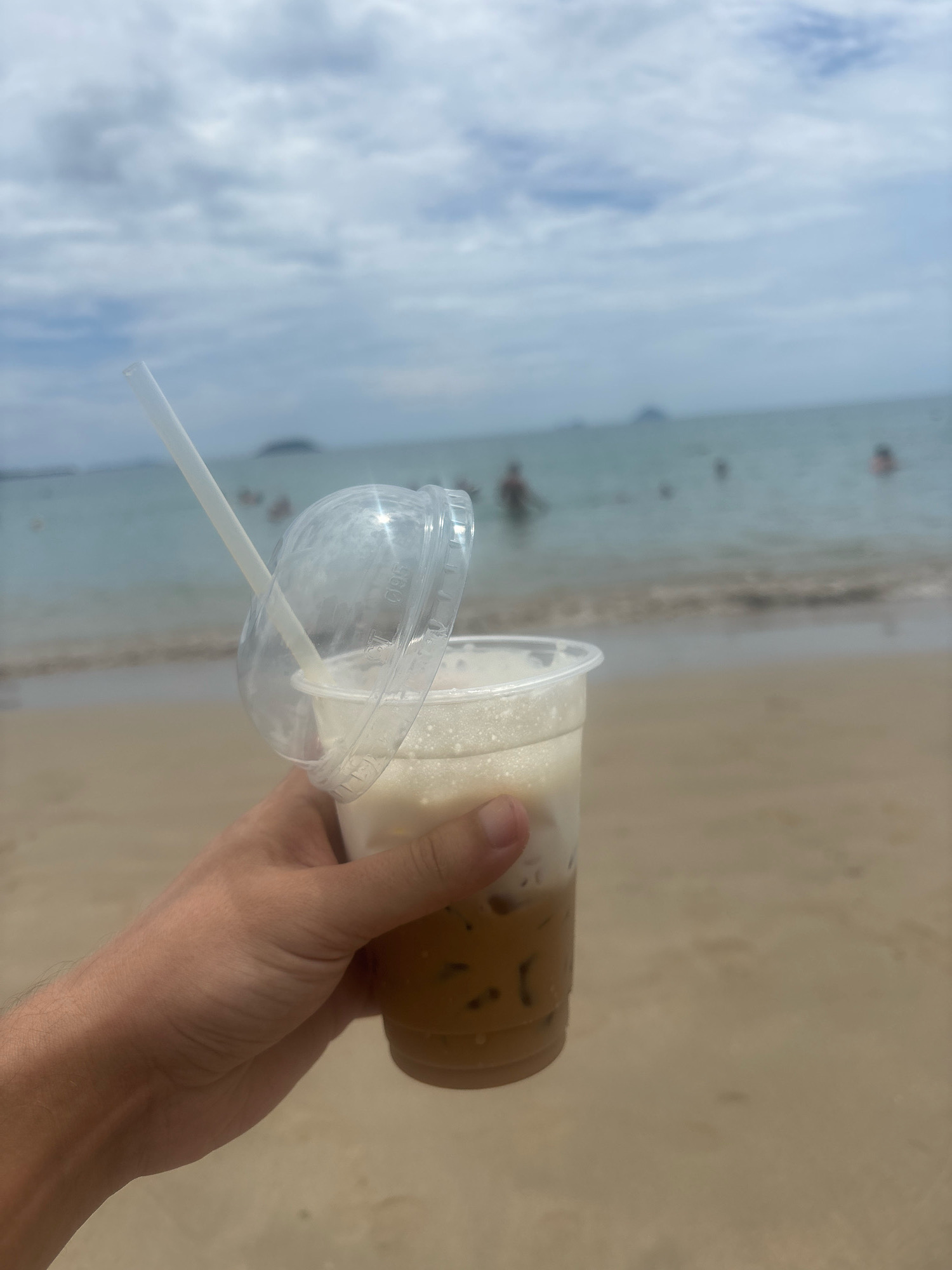 A close-up shot shows a person's hand holding a clear plastic cup with an iced beverage and a white straw. The drink is layered with a dark brown liquid at the bottom, ice cubes, and a foamy white top. A clear plastic lid is ajar, allowing the straw to extend upwards. The background is a beach scene with the ocean and a cloudy sky. Several blurry figures are visible in the water, some appearing to be swimming. The sand is a light brown color. The scene suggests a relaxed day at the beach in Nha Trang, Vietnam.