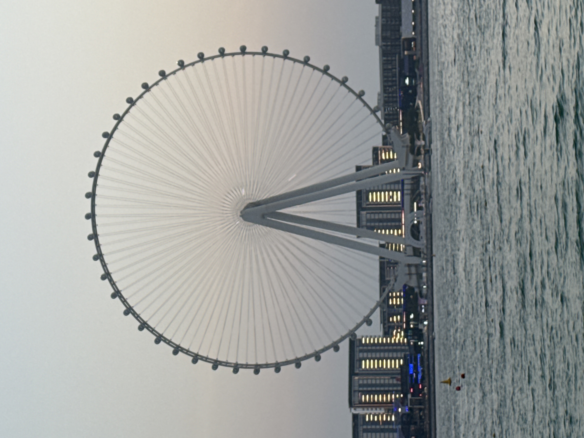 The image depicts the Ain Dubai observation wheel, a prominent landmark in Dubai, United Arab Emirates. The wheel is a massive structure, with its characteristic spokes and circular frame dominating the upper half of the image. It stands on a broad base with two angled support legs. In the background, several modern high-rise buildings with illuminated windows are visible, suggesting an urban setting. The scene is set against a pale, overcast sky, possibly indicating twilight or a hazy day. In the foreground, a body of water, likely the Persian Gulf, is visible with gentle ripples on its surface. Several buoys, one yellow and two red, are floating in the water. The overall impression is one of a serene, yet grand, city skyline with the iconic observation wheel as the focal point.