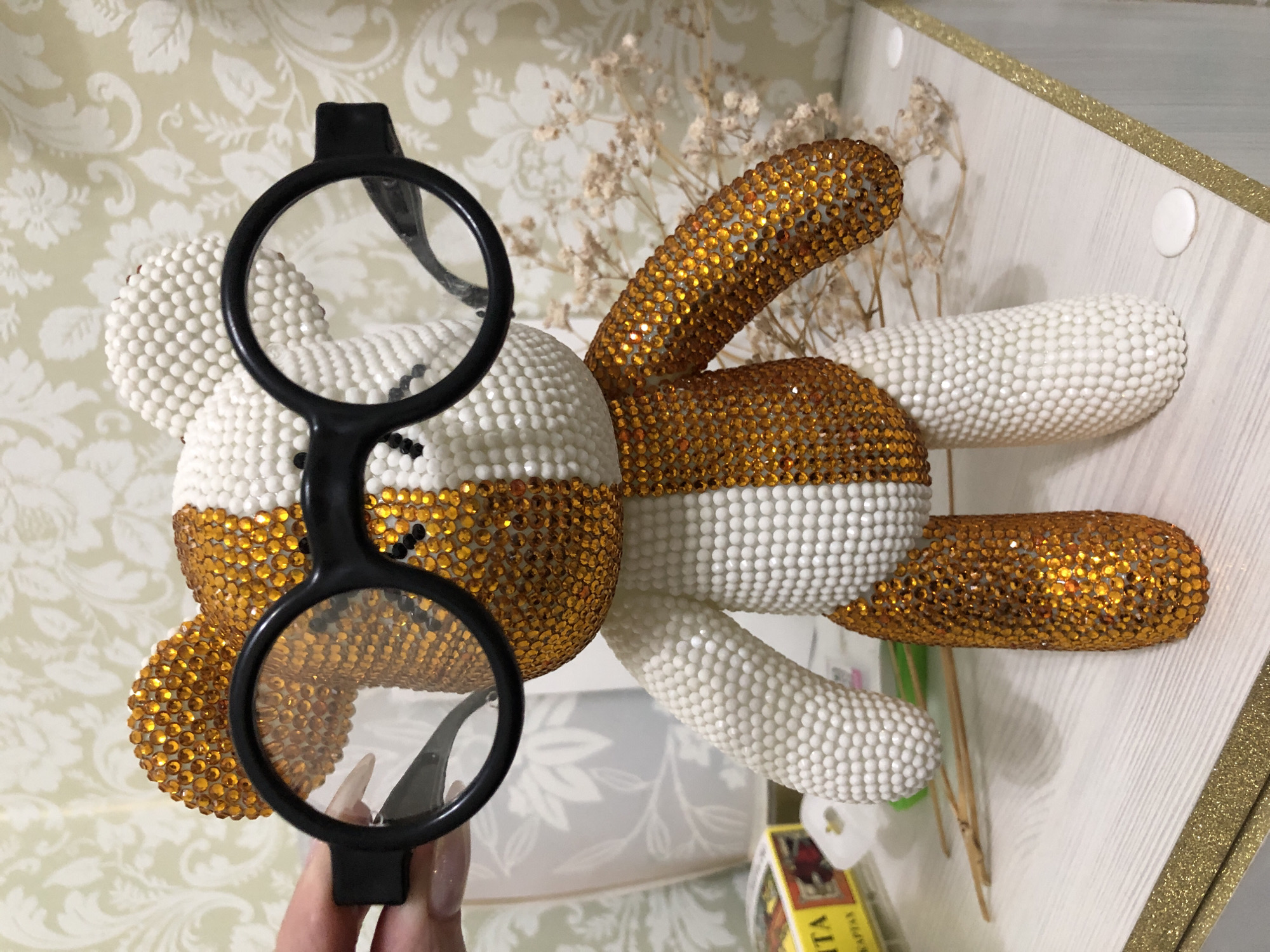 A photograph shows a beaded toy bunny lying on its side. The bunny is decorated with gold and white beads. A pair of black-rimmed eyeglasses is held in front of the bunny’s face. The toy is positioned inside a box with a floral patterned background. A hand holds the eyeglasses. Some yellow items, possibly boxes, are at the bottom of the image. The location is not discernible from the provided image.