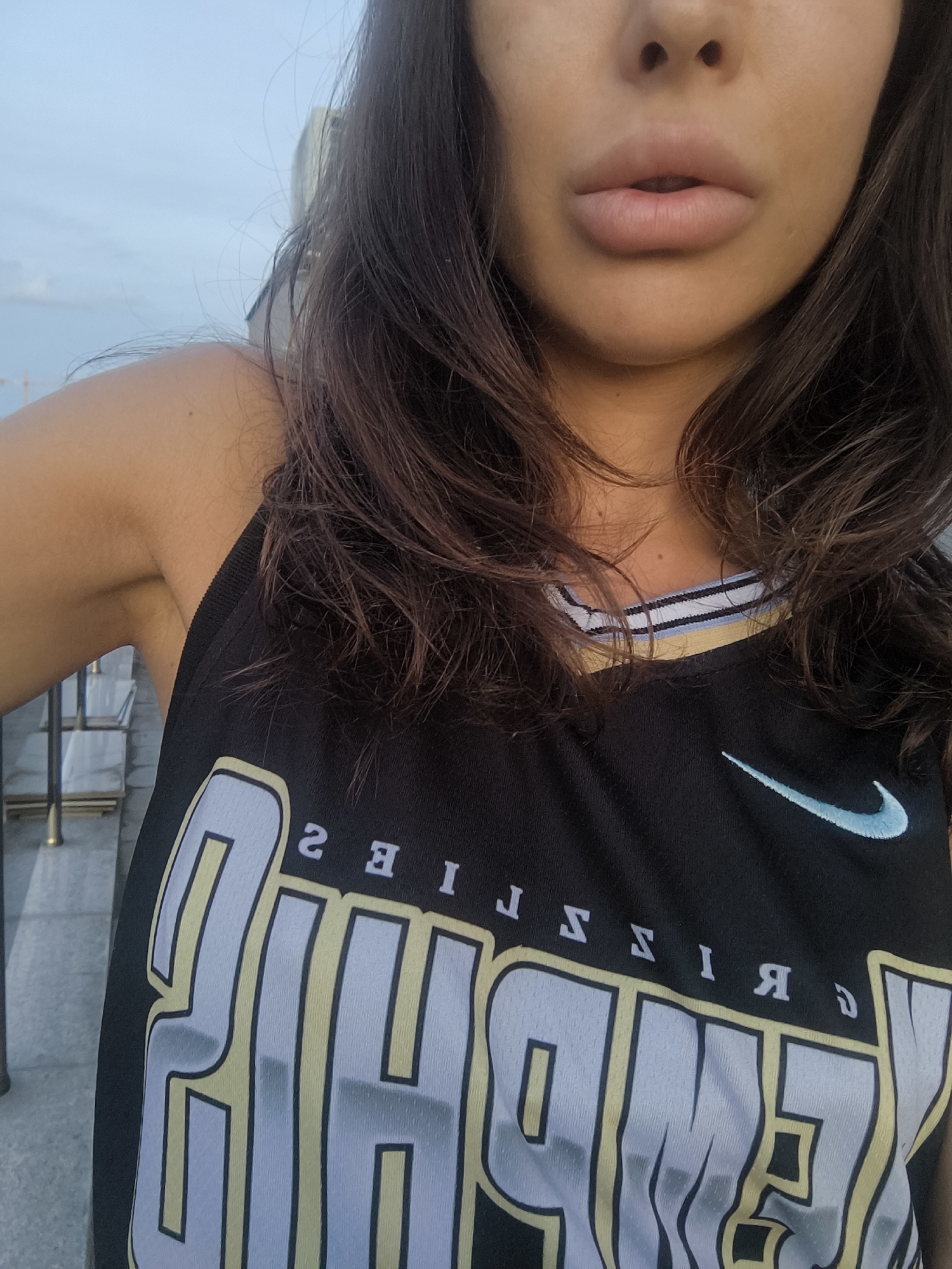 The image shows a close-up of a woman's upper torso and face. She has dark hair and light-colored lips. She is wearing a black tank top with yellow and white lettering and a light blue Nike logo. The background appears to be an outdoor setting with a cloudy sky and some buildings visible in the distance. The overall impression is that this is a selfie taken outdoors during the day. There is no information in the image to suggest this location is Sofia, Bulgaria.