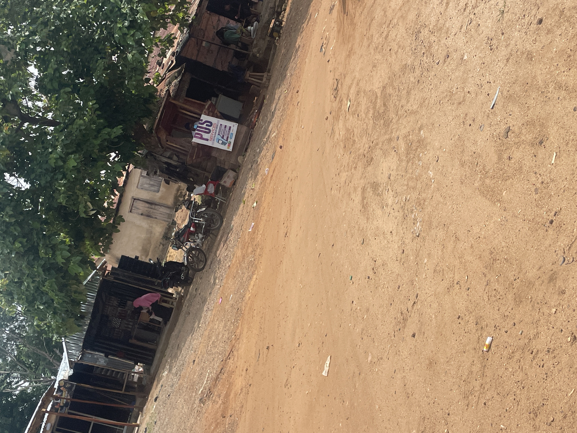 The image depicts an outdoor scene in Lokoja, Nigeria, characterized by a dirt road and several small, informal shops or stalls. A large tree with lush green foliage dominates the upper portion of the frame, casting shade. In the foreground, the dirt road is uneven and strewn with small debris.
To the left, a row of structures with corrugated iron roofs and open fronts suggests small businesses. One stall has a person wearing a pink headscarf visible inside, seated behind a counter. Two motorcycles are parked in front of these stalls.
Further to the right, another cluster of similar structures is present. A prominent sign with the text "POS SERVICES AVAILABLE HERE" is displayed, indicating a financial service point. A person is seen standing near this sign, and another individual is seated within one of the stalls. A small generator is visible next to one of the buildings. The general environment appears to be a local marketplace or a street with informal commercial activity.