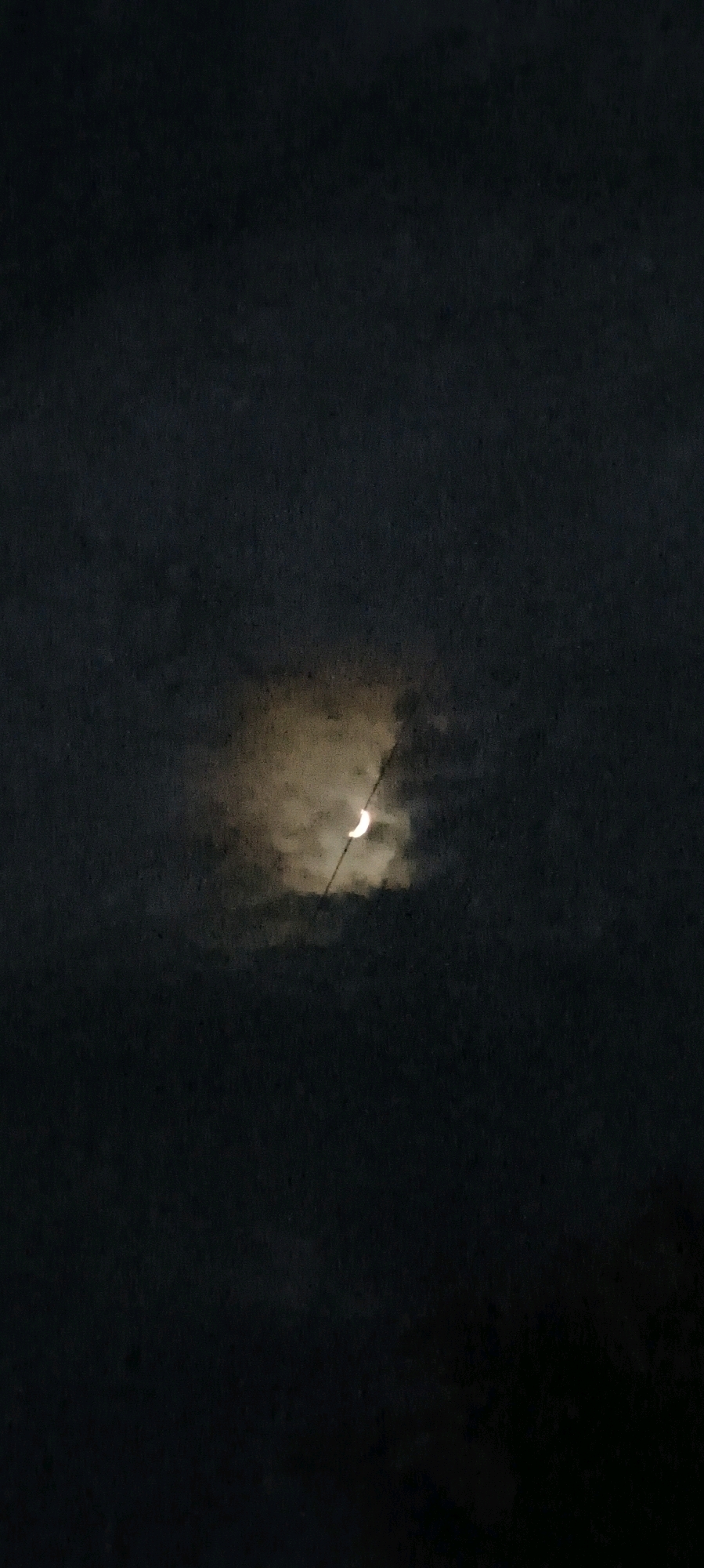 The image depicts a dark, cloudy night sky over Bansarola, India. A crescent moon is partially visible through a break in the clouds, illuminated by an unseen light source. A thin, dark line, possibly a wire or branch, crosses the frame in front of the moon and clouds. The overall atmosphere is dim and overcast.
