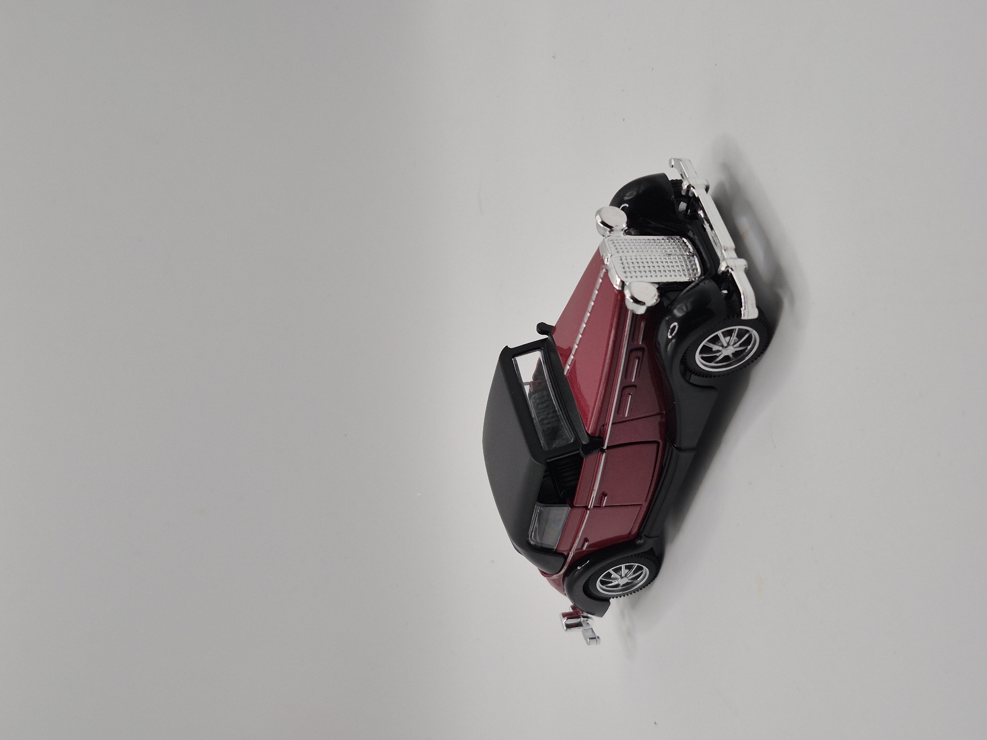 A maroon and black toy car is positioned on a plain, light gray surface. The car is a vintage convertible with a detailed silver grille, headlights, and bumper. It has black fenders and a black convertible top. The wheels are silver with black tires. The car is viewed from a slightly elevated, three-quarter front angle. There are no people or any indication of a specific location within Schwäbisch Gmünd, Germany, in this image.