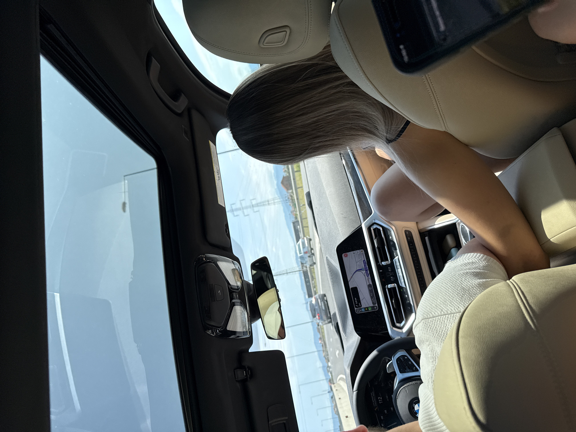 The image shows the interior of a car with a sunroof open. The perspective is from the back seat, looking forward. A person's arm is resting on the passenger seat, and a woman with blonde hair is visible in the passenger seat. She is wearing a black bikini top. The dashboard of the car is visible, with a navigation screen displaying a map. The car is on a highway, with other cars in the distance. The sky is blue with some clouds. There are power lines and buildings visible outside the car. The location is identified as Sanpetru, Romania.