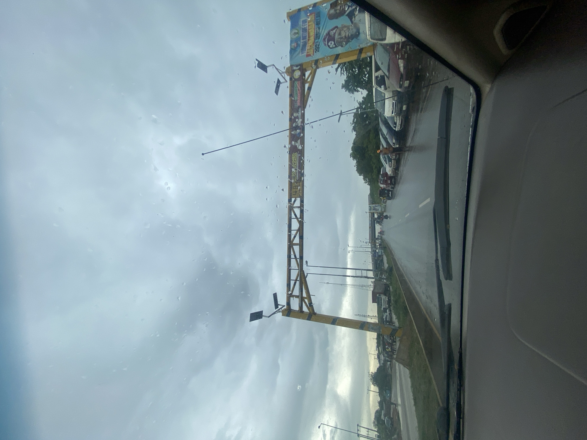 The image captures a view from inside a vehicle looking through a rain-streaked windshield towards a busy road in Keffi, Nigeria. The sky is overcast with heavy clouds, and the road surface is wet, indicating recent or ongoing rainfall. A large yellow overhead structure, possibly a toll gate or an archway, spans the road, decorated with lights and a prominent banner on the right side featuring portraits of political figures and campaign slogans. Several vehicles are queued on the road, suggesting traffic congestion. A few individuals are visible near the roadside and on the median. In the foreground, the dashboard and wipers of the vehicle are visible, framing the scene.