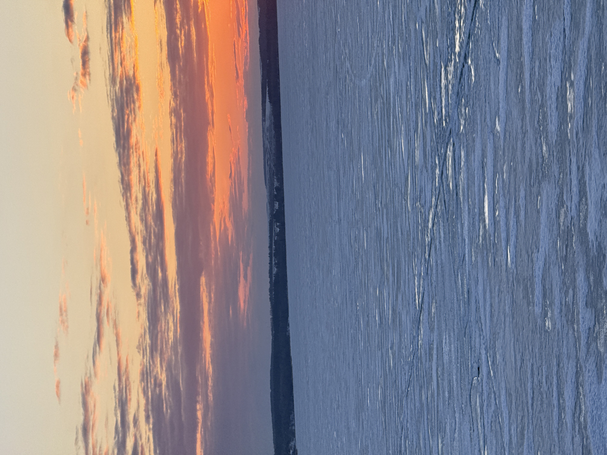 The image depicts a landscape scene with a dramatic sky on the left and a body of water on the right, potentially in Votkinsk, Russia.
**Sky:** The sky exhibits vibrant colors, suggestive of a sunrise or sunset. Hues of orange, pink, and gray are present within the cloud formations.
**Water:** The body of water, possibly a lake or river, has a blue-gray tone, appearing somewhat rough with visible streaks or patterns on its surface.
**Land:** A dark, thin strip of land or coastline is visible between the sky and water, potentially indicating the opposite shore.
Without specific identifying features or landmarks, a definite confirmation of the location being Votkinsk is uncertain. However, the general characteristics of the scene (sky, water, distant land) are consistent with a view over a body of water at dusk or dawn in a northern region.