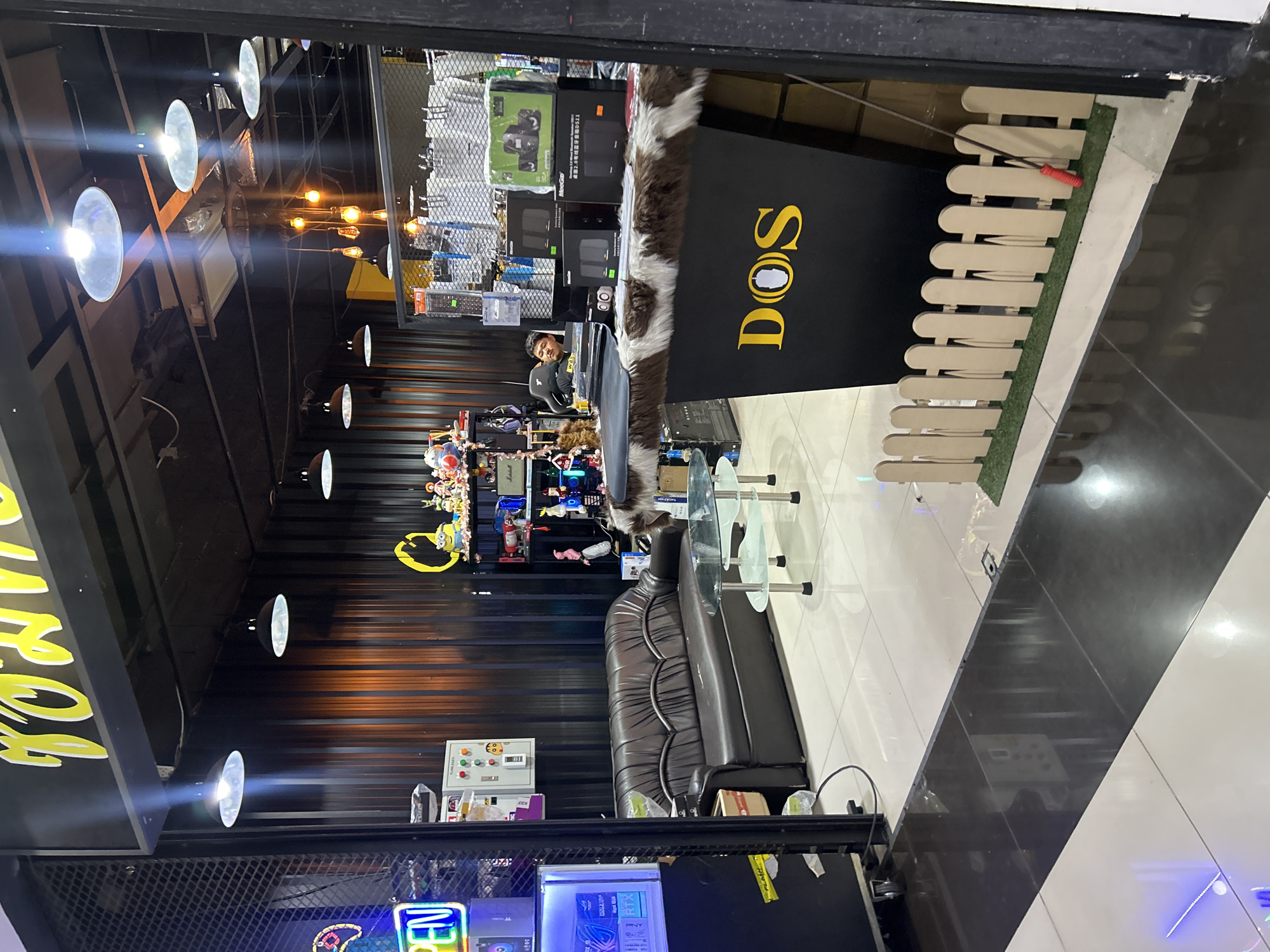 The image shows the interior of a store located in Khlong Luang Town Municipality, Thailand. The store appears to sell electronics and possibly computer accessories, judging by the visible products like computer speakers and graphics cards. A dark brown leather sofa is positioned in the foreground, with a glass coffee table in front of it. Behind the sofa, shelves are stocked with various items, including figurines and what appear to be electronics.
A counter with a furry, cow-print trim is prominent in the center of the image. A man is seated behind this counter, looking towards the right. The counter itself has the logo "DOS" displayed in large yellow lettering, with a stylized computer mouse incorporated into the lettering. In front of the counter, a small, decorative picket fence with artificial grass is placed on the floor.
The store's interior features dark corrugated metal walls and a black ceiling with multiple light fixtures. A neon "OPEN" sign is visible on the left side of the image. The floor is tiled with a combination of white and dark reflective tiles. The overall impression is that of a retail space with a somewhat eclectic and modern aesthetic.