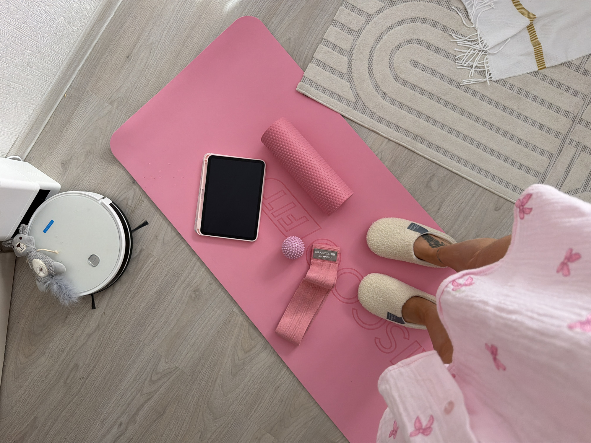 This is a top-down, eye-level shot of a pink yoga mat laid out on a light wood-effect floor. The mat is adorned with a subtle embossed "FIT" logo. On the mat, from top left to bottom right, are: a tablet in a pink case, a pink resistance band, a small pink massage ball, and a pink foam roller. A person's feet, wearing fluffy cream slippers, are visible at the bottom of the mat. To the right of the yoga mat is a light-colored rug with a geometric pattern of grey arched lines and a white fringed throw draped on its corner. In the upper left of the image, a white robot vacuum cleaner is present, topped with a gray plush toy. The background consists of a white wall with a subtle texture and a white baseboard. This scene is not specific to Moscow, Russia, as there are no identifiable landmarks or contextual elements suggesting that location.