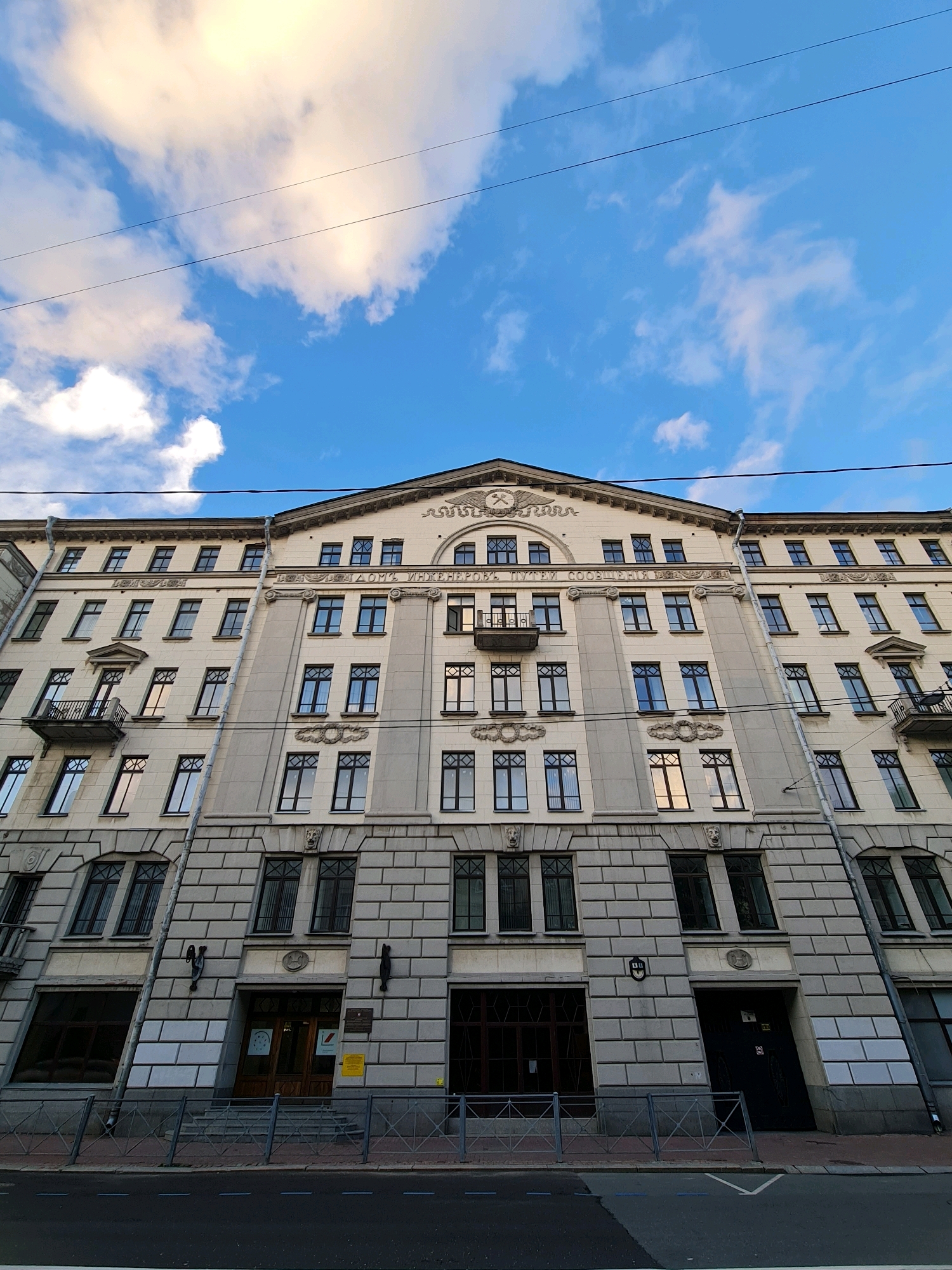 This image depicts the facade of the "House of Engineers" (Дом инженеров) located at 19 Nevsky Prospekt in Saint Petersburg, Russia. The building is a multi-story structure with classical architectural elements, characterized by its beige and grey stone facade, numerous windows with dark frames, and decorative detailing. The perspective is from a low angle, looking up towards the sky. Above the building, a bright blue sky is visible with scattered white and yellow clouds, illuminated by sunlight. Two thin, dark wires run horizontally across the upper portion of the frame, indicating the presence of overhead electrical or communication lines. The ground level of the building features several entrances, staircases, and a street with a sidewalk and road markings. The scene appears to be devoid of any visible people or ongoing activity.