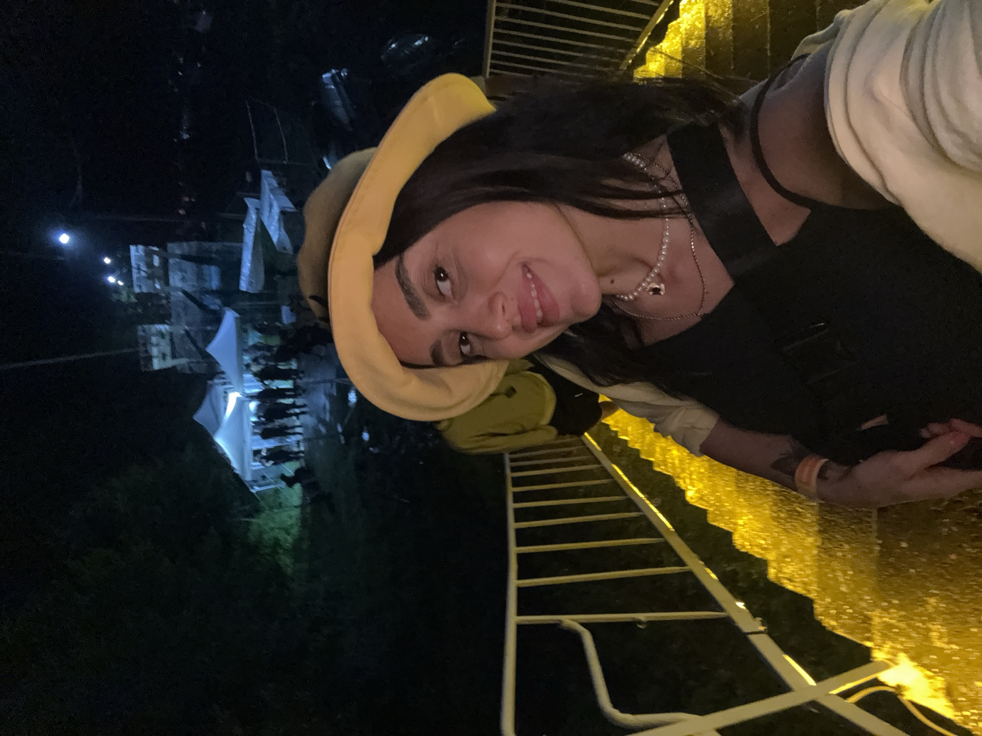 A nighttime, slightly elevated, wide-angle selfie shows a woman with dark hair, wearing a yellow bucket hat, a black top, and a pearl necklace, smiling at the camera. She is standing on a brightly lit yellow staircase, with a white railing on her left. Behind her, a person in a yellow hoodie and black shorts is walking up the stairs. In the background, several white tents are set up, illuminated from within, and a group of people are gathered. Beyond the tents, a stone structure, possibly part of a historical building, is visible. The overall environment suggests an outdoor event or festival taking place at night in Poti, Georgia, potentially near a historical site.