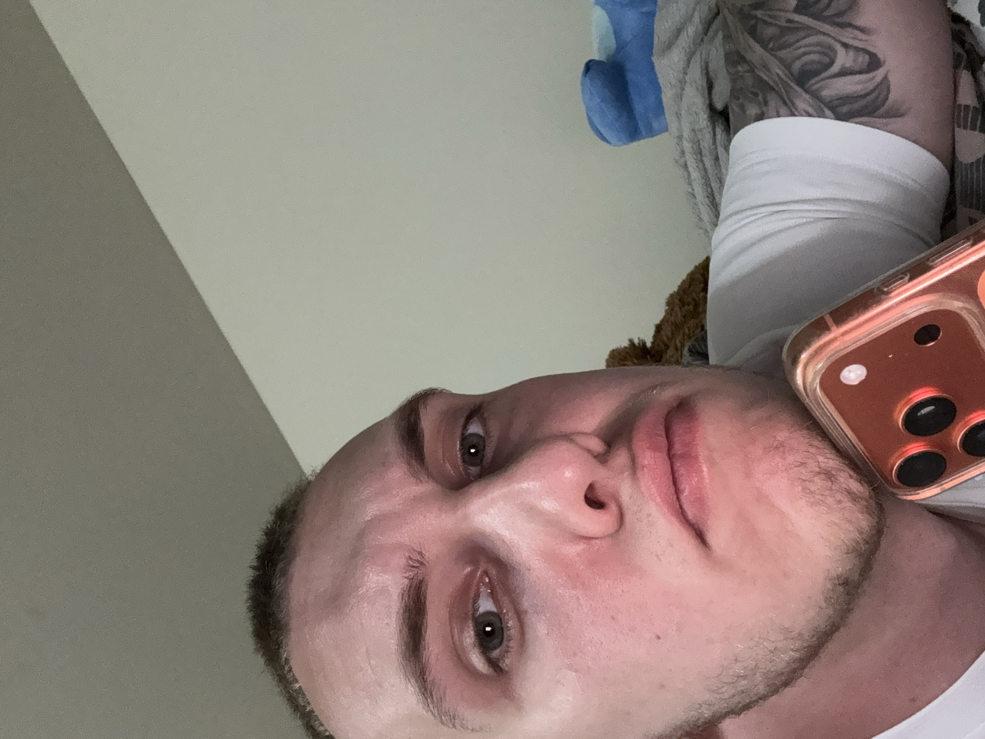 An extreme close-up, low-angle shot captures a male individual indoors, likely in Dobrogled, Bulgaria. The person is fair-skinned with light stubble on his chin and jawline, and light-colored eyes that gaze towards the viewer. His face exhibits some redness, particularly around the eyes and cheeks. He appears to be lying down, with the camera positioned below him, creating an upward perspective.
He is wearing a white t-shirt or undershirt. A detailed black and white tattoo, possibly part of a sleeve, is visible on his upper left arm/shoulder area. To the right of the frame, a smartphone with a distinctive rose gold or copper-colored case is visible, showing its camera lenses.
The background primarily consists of a plain light off-white or light grey wall, with a diagonal shadow dividing a section of it. Other elements in the background include a soft blue fabric object (possibly a blanket or pillow) and a textured brown fabric, suggesting a casual, comfortable setting such as a bedroom. There is no visible text. The lighting suggests an indoor setting, likely artificial. The person's expression is neutral, bordering on contemplative or relaxed.