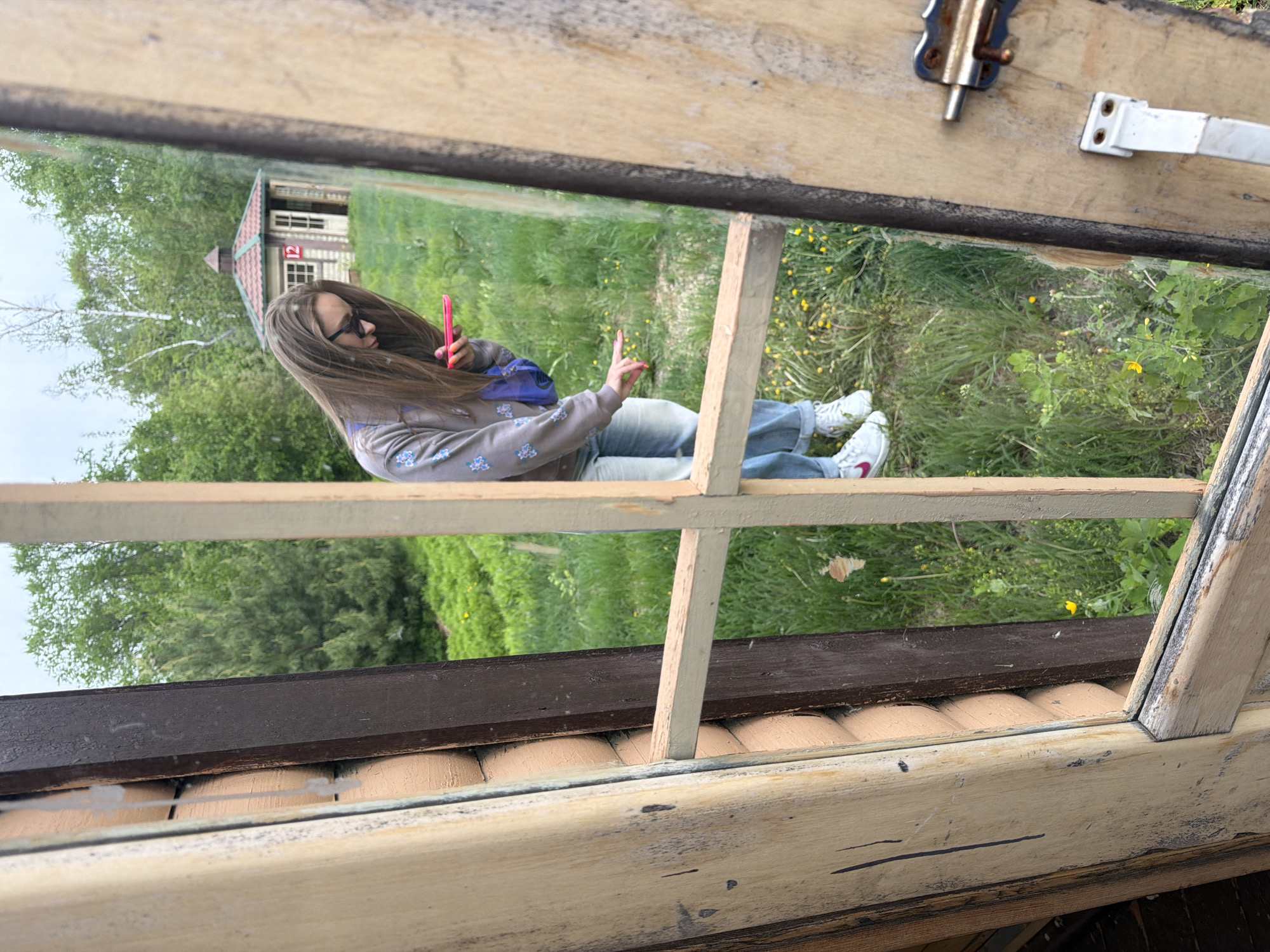 A woman is seen through an old window, likely taking a selfie with a pink phone. She is wearing sunglasses, a patterned sweatshirt, jeans, and white sneakers. The background outside the window includes green grass, trees, and a small house with a red roof. The window frame is made of weathered wood, with flaking paint. I cannot confirm the location of this scene is in Birobidzhan, Russia, as there is no evidence in the image to confirm the location.