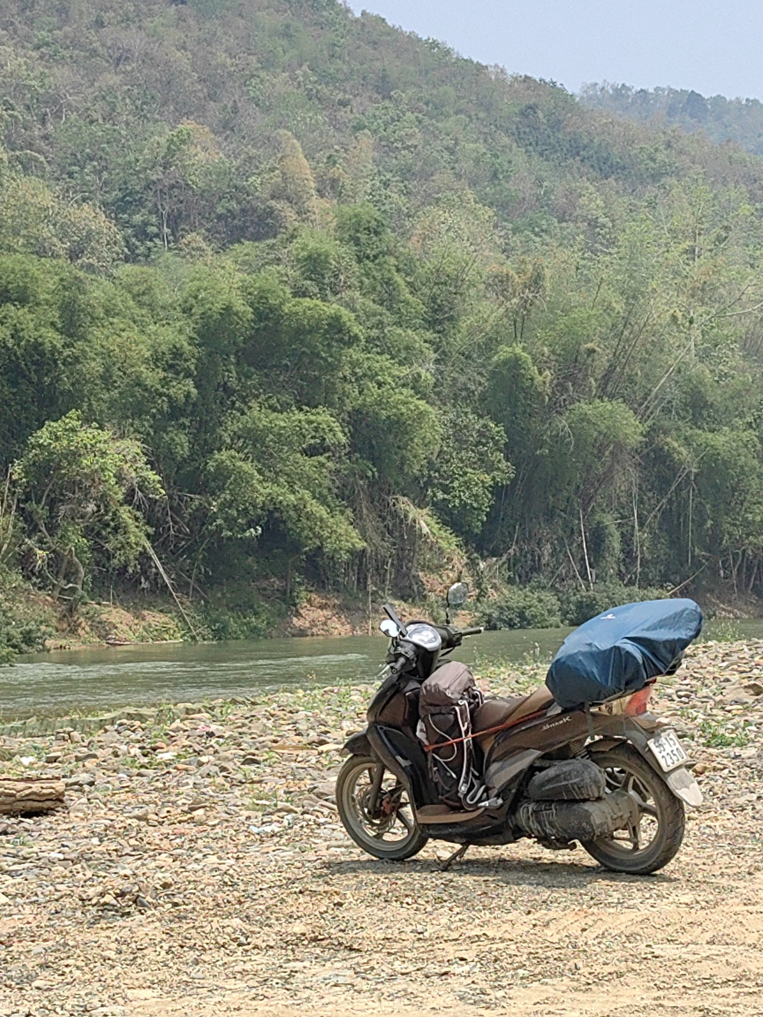 A motor scooter with luggage is parked on a rocky shore next to a river. The scooter is dark-colored, with bags strapped to the front and a large blue bag secured on the back. A river flows parallel to the shore, reflecting the sky and surrounding vegetation. The riverbank consists of light-colored pebbles and sand. Lush green trees and vegetation cover the hillside in the background. Xã Cô Ba, Vietnam.