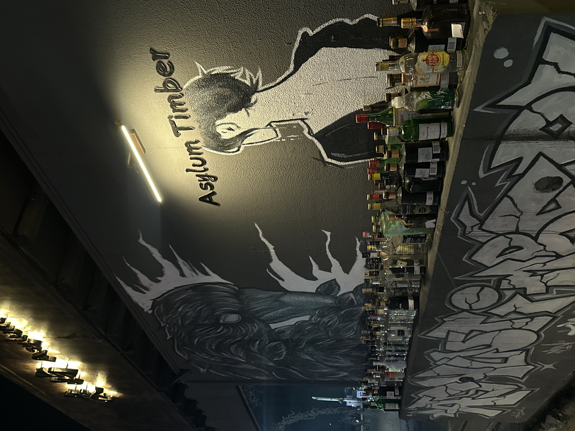 The image depicts an exterior view of a bar or establishment at night. The wall behind a row of liquor bottles features a large mural. On the left side of the mural is a stylized drawing of a creature with long, flowing hair or plumes. To the right of the creature is a drawing of a person's head and shoulders, facing away from the viewer, with a black outline and white fill. Above the mural, the words "Asylum Timber" are visible in raised metal lettering. The establishment is identified as being in the Chongqing High-tech Zone, China. A strip of illuminated signage is mounted above the wall, casting a warm glow. The row of bottles suggests a bar setting, with various types and brands of liquor on display. The foreground is dominated by graffiti art on a concrete barrier. The overall lighting is dim, with accent lighting from the signage and a strip light illuminating the "Asylum Timber" text. No people are visible in the scene.