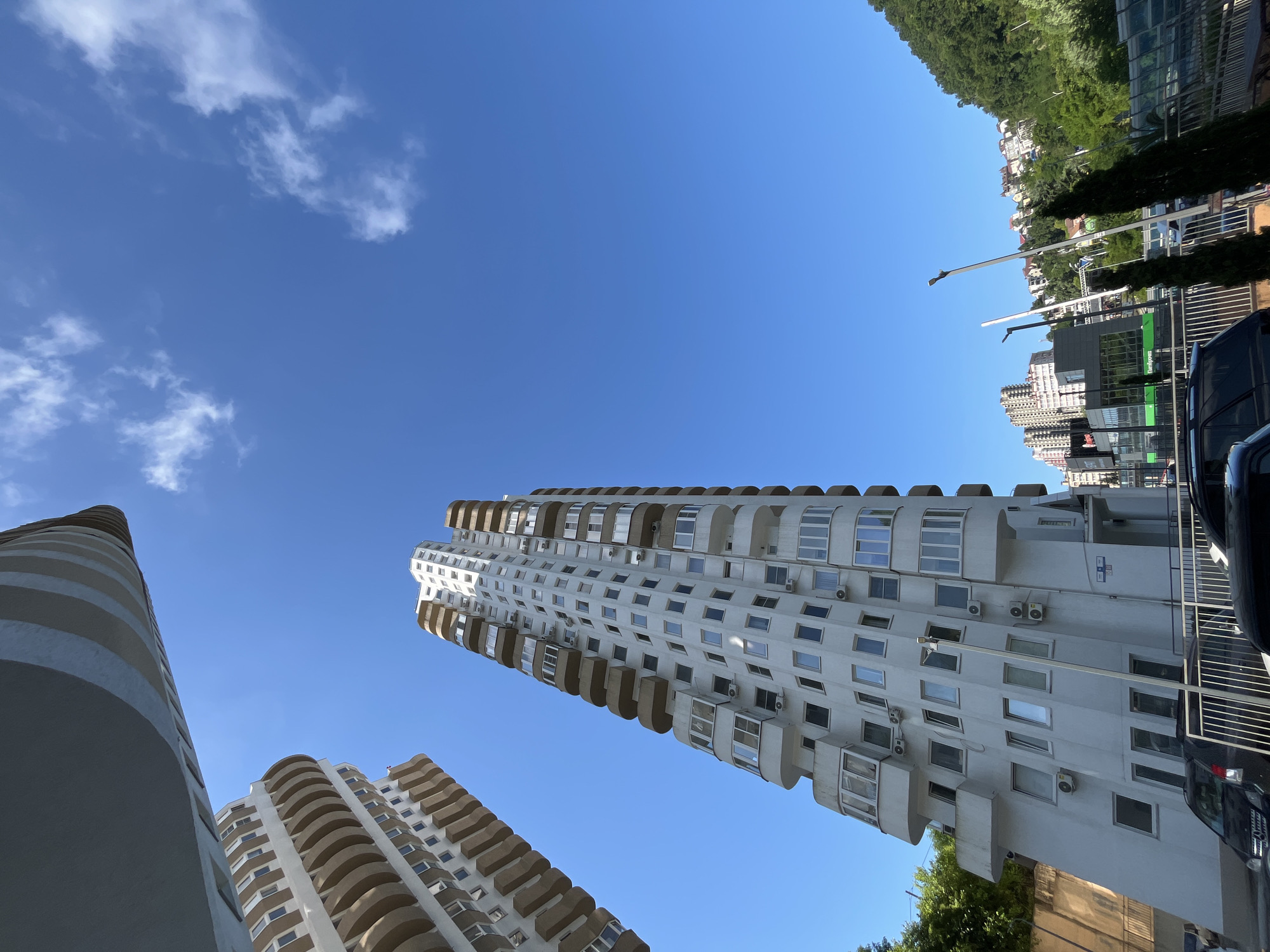 This image captures a low-angle view of modern apartment buildings in Sochi, Russia, under a clear blue sky with sparse white clouds. In the foreground, a tall, white apartment building with numerous balconies and windows dominates the frame, leaning slightly to the right as if viewed from a very close perspective. To its left, another similar building is partially visible, also featuring curved balconies. The buildings have a contemporary architectural style, characterized by their height, white facades, and balconies.
In the background, other residential buildings and a commercial structure with signage are visible on a hillside covered in lush green trees. The overall impression is one of a developed urban area integrated with a natural, hilly landscape, typical of Sochi's coastal environment. The image appears to be taken during daylight, with bright sunlight illuminating the scene. There are no people clearly visible in the image, and the focus is on the architecture and the sky. The presence of parked cars and a fence in the immediate foreground suggests a street or parking area.