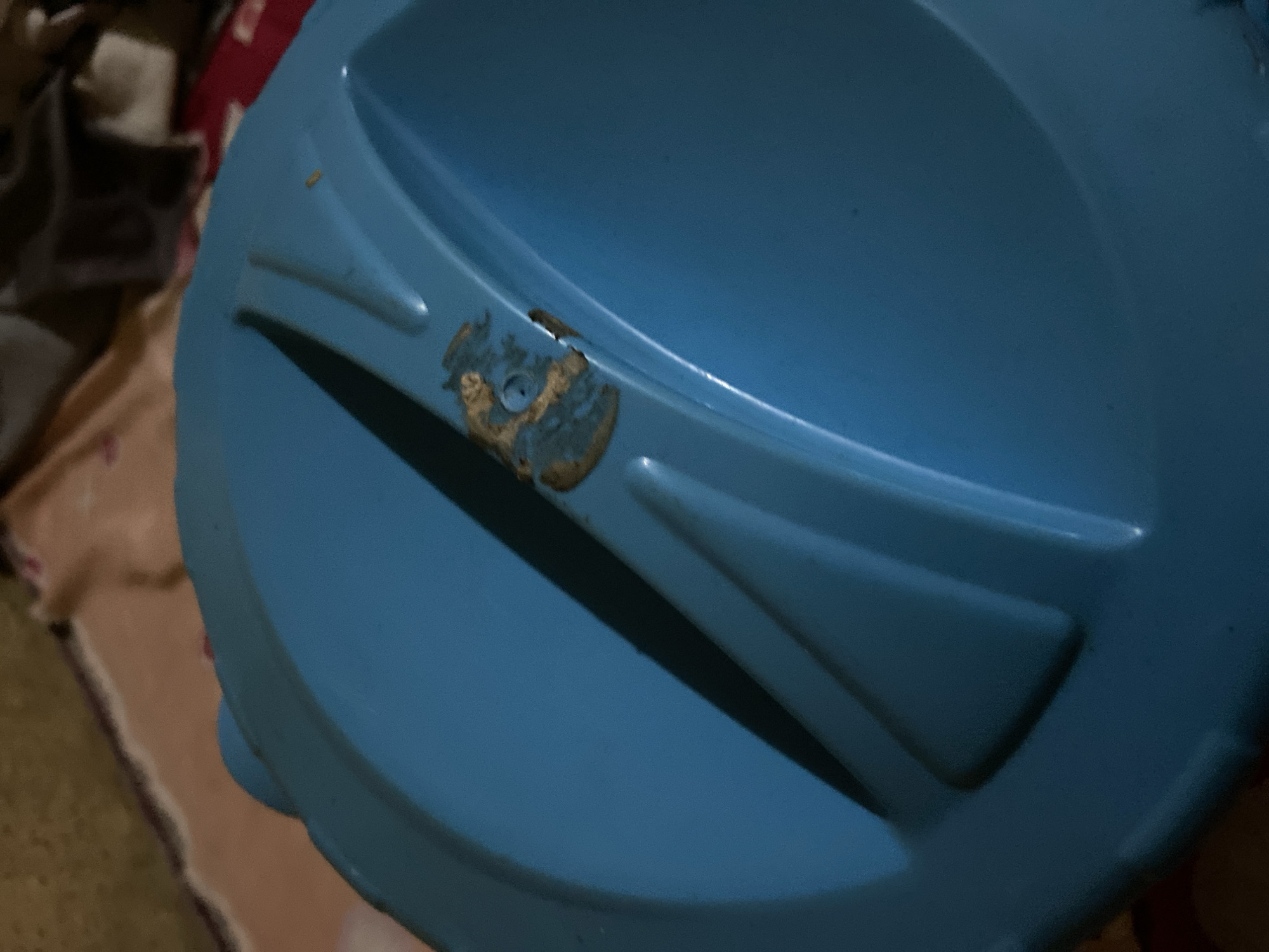 A close-up, overhead shot shows a blue plastic lid with a textured surface. The lid is angled, with one side closer to the camera. A patch of what appears to be damaged or worn material is visible on the lid, revealing a lighter color underneath. The background is out of focus and appears to be a patterned fabric, possibly a bedspread or blanket, in muted colors of beige, brown, and red. The overall lighting is dim. The scene is not identifiable as being in Jalingo, Nigeria, or any specific urban environment.