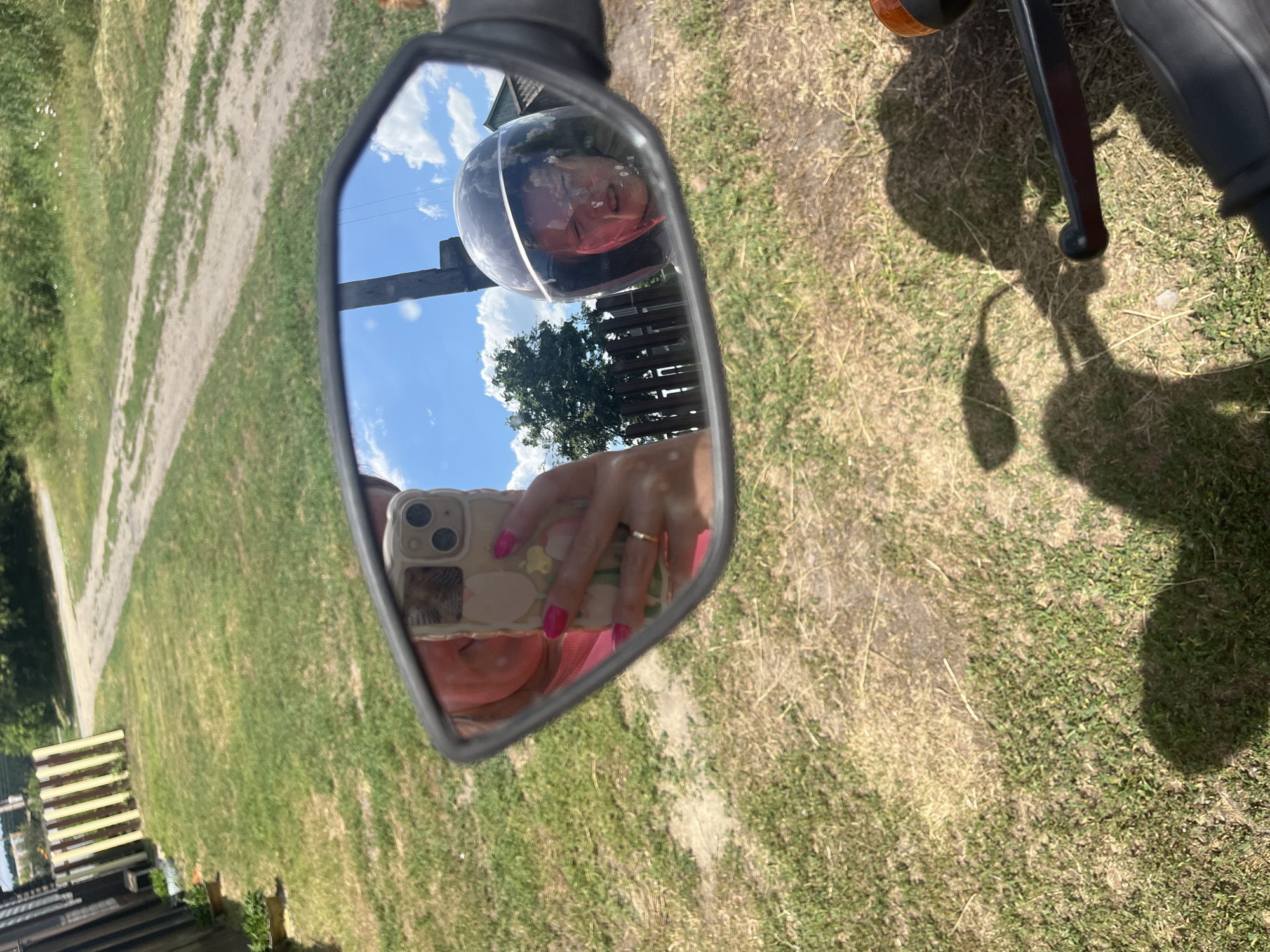 The image is a first-person perspective taken from a motorcycle, reflecting a rearview mirror. In the mirror, a person is visible holding a smartphone, taking a selfie. The person has bright pink fingernails and is wearing a pink top. To the right of the selfie, a child wearing a dark helmet with a clear visor is reflected, with their face visible through the visor. The child appears to be grimacing.
The background reflected in the mirror shows a dirt road with grass and trees on either side, suggesting a rural or suburban setting. There is a wooden fence and a building visible in the distance. The sky is blue with scattered white clouds. The foreground of the image, outside the mirror, shows the motorcycle's handlebars and a shadow cast on the dry, grassy ground, indicating a sunny day.
The location is identified as Сухринское, Russia.