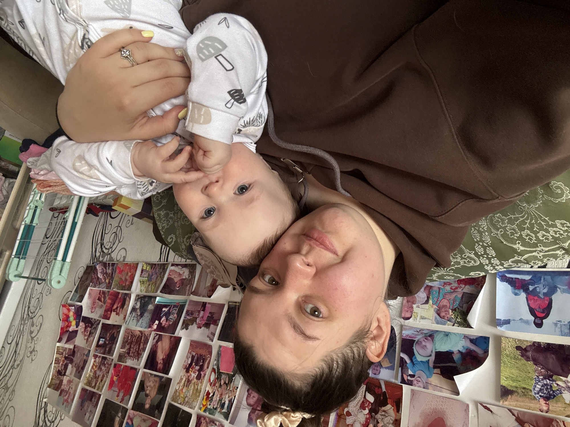A woman and baby are lying together in what appears to be an indoor setting. The woman has fair skin, brown hair pulled back in a scrunchie, and is wearing a brown hoodie. The baby has fair skin, blue eyes, and is wearing a white onesie with a mushroom pattern. The woman is looking at the camera. The location cannot be determined, though it is purportedly in Lukintsy, Russia. Behind them is a wall covered in photographs. A garment rack is visible on the left.