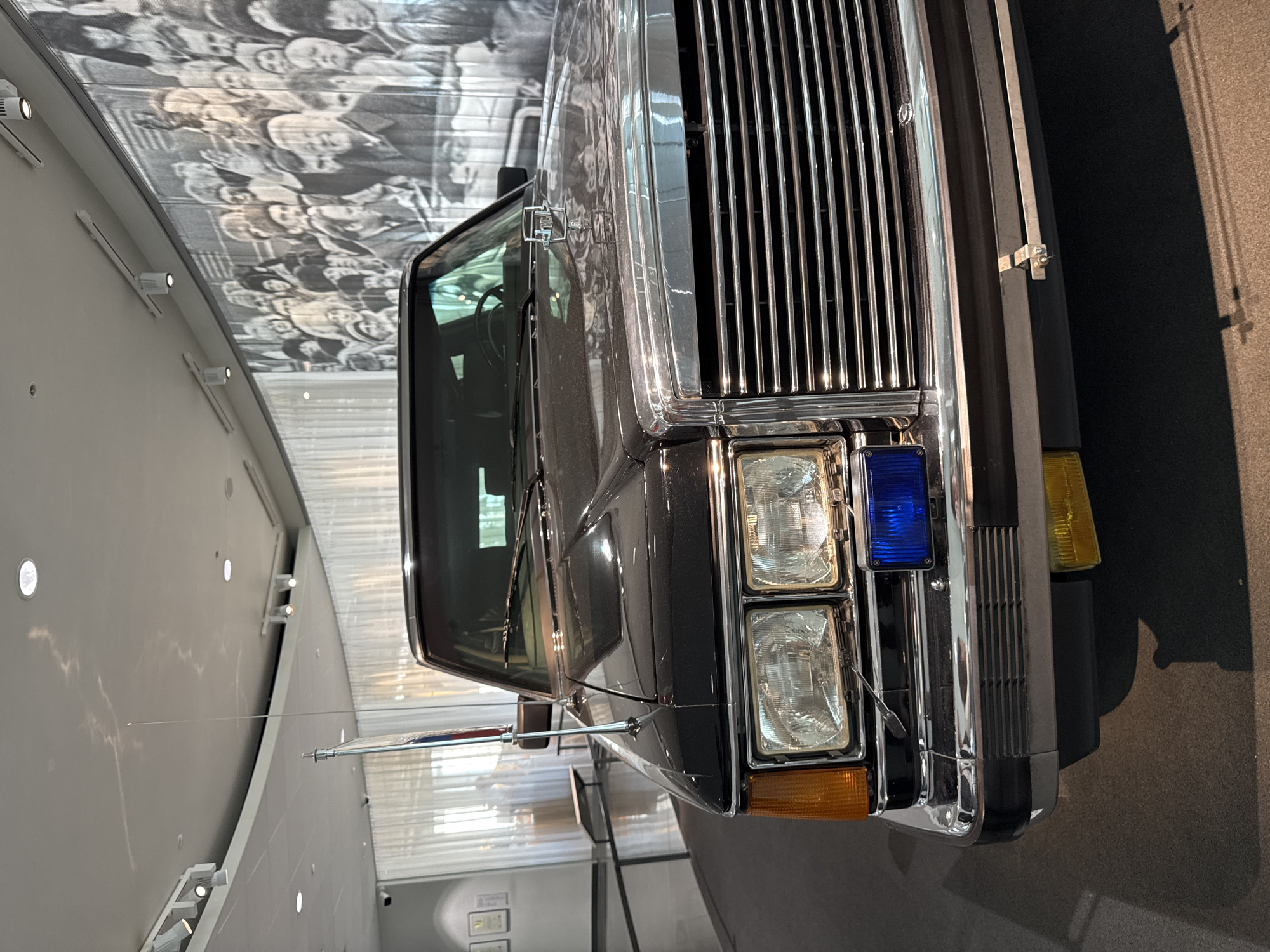 The image captures the front of a black car with chrome accents and a distinctive vertical grill, a square headlight configuration (two headlights on each side), and a small antenna extending horizontally from the side. The scene appears to be inside a museum or exhibition hall, given the lighting and the presence of a large photo mural on the ceiling, which features an indistinct crowd of people in black and white. The car is resting on a dark surface, and the background also includes a light-colored wall with some display items. This is Yeltsin Center in Yekaterinburg, Russia.