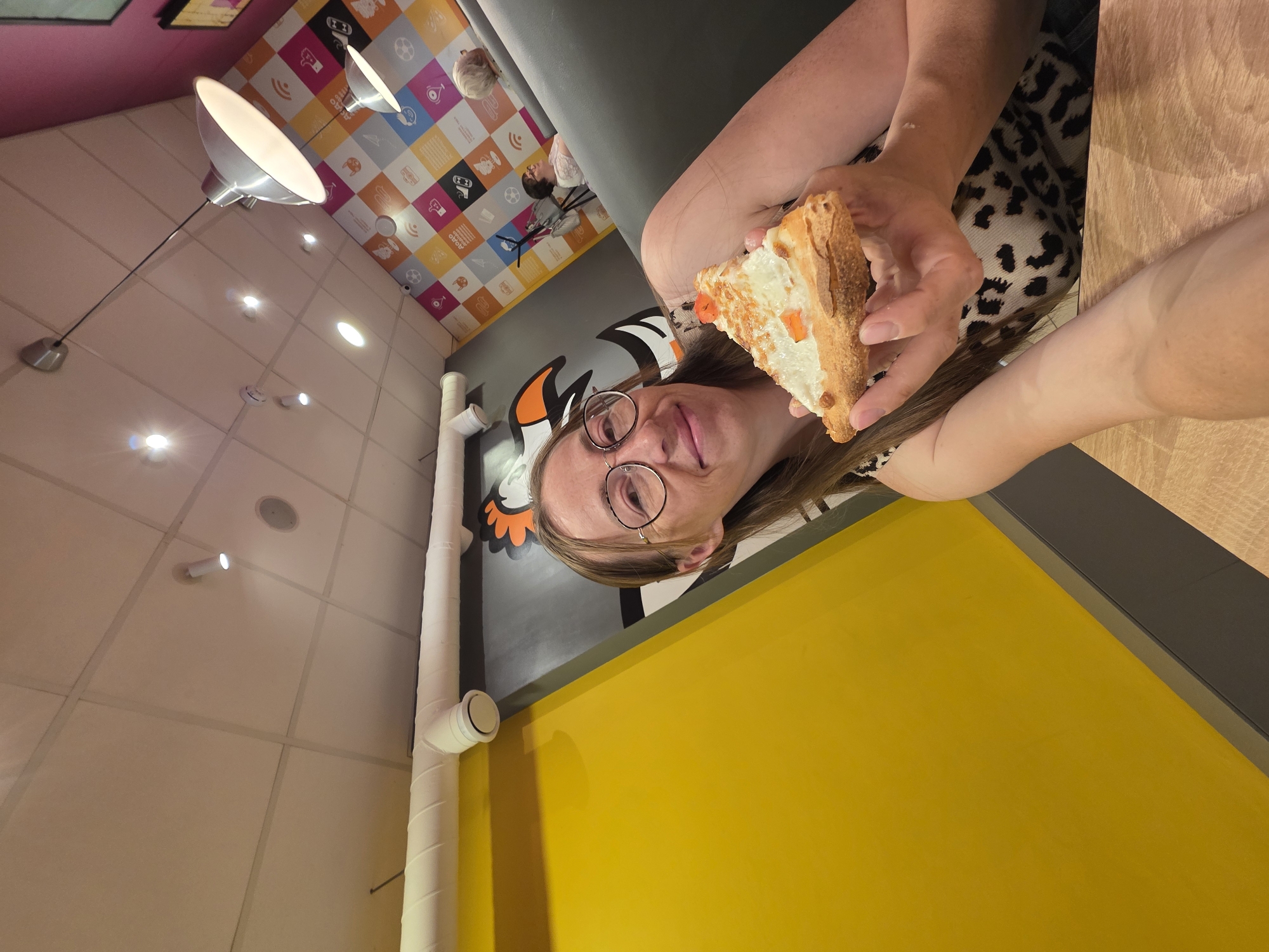 A person wearing glasses is holding a slice of food, potentially a savory tart or pizza, in a room with colorful walls and ceiling. The interior features a combination of yellow, gray, white, and multicolored patterned surfaces. Overhead lighting fixtures are visible, including a large, dome-shaped lamp extending from the ceiling. Piping runs along the wall. The exact location within the city of Chaikovsky, Russia, cannot be determined from the image alone.
