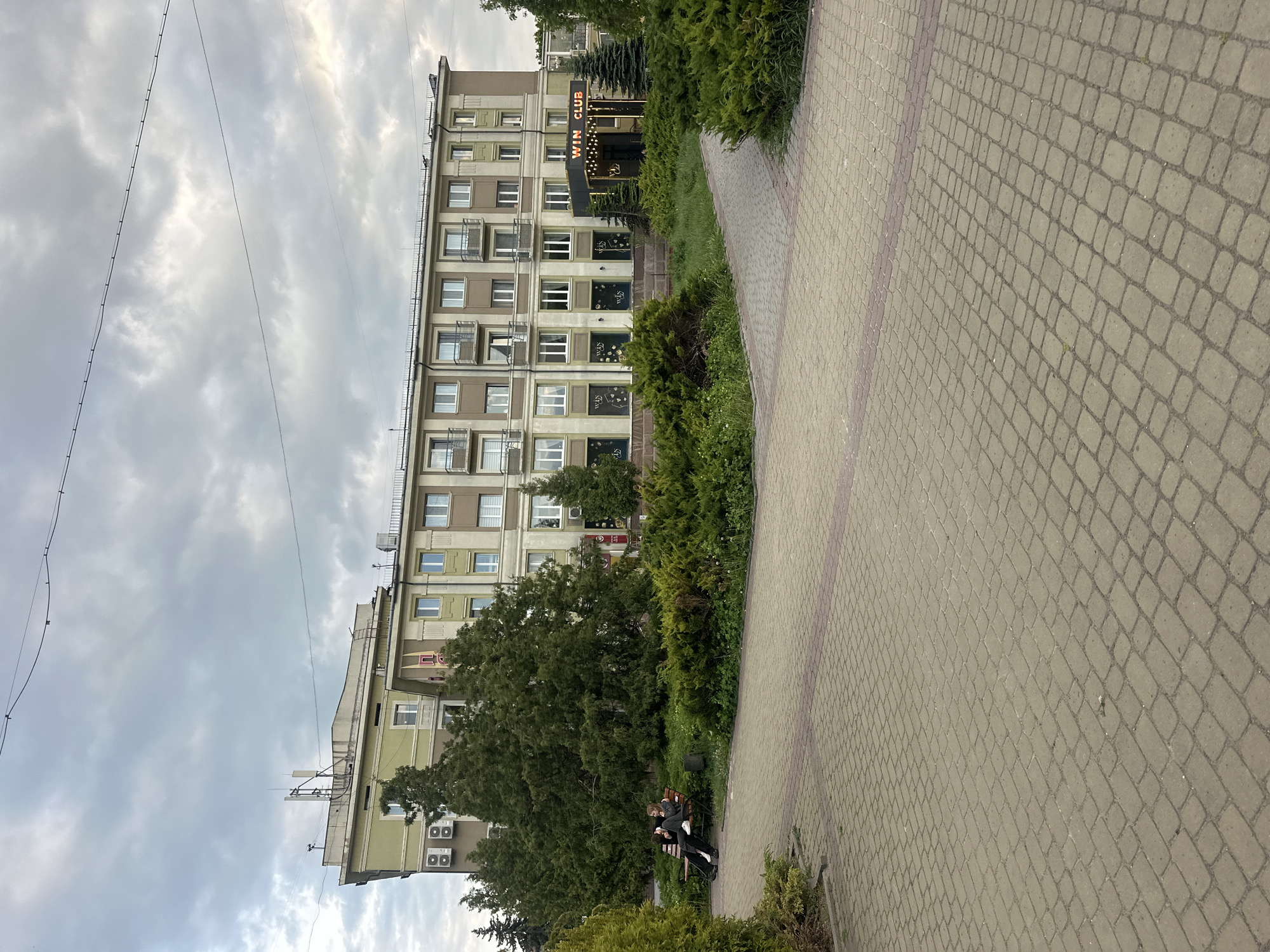 The image depicts an urban landscape in Ternopil, Ukraine. A multi-story building dominates the left side of the frame, with a "WIN CLUB" sign visible above the entrance. The building's facade is characterized by numerous windows and balconies. A paved walkway stretches across the right side of the image, constructed from small, rectangular paving stones. Along the walkway, shrubs and trees are present. Two people are seated on a park bench near the bottom of the image, their actions unclear. Overhead, power lines are visible against a cloudy sky.