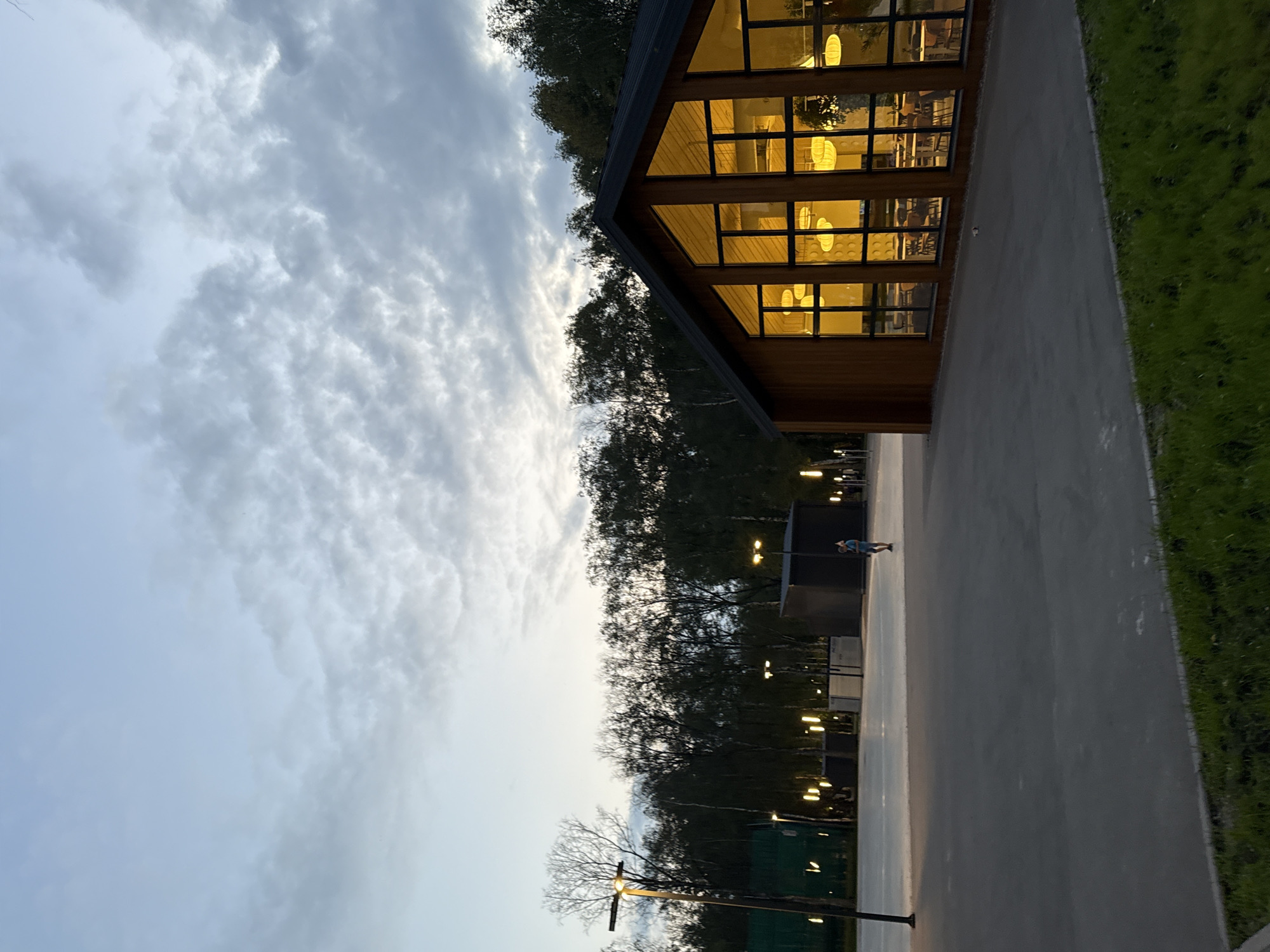 The image depicts an outdoor scene at dusk, with a modern wooden building with large windows prominently featured on the right. The building's interior is illuminated with warm yellow light, revealing tables and chairs within, suggesting it is a cafe or restaurant. In the background, a line of trees forms a dark silhouette against the twilight sky, which is partly cloudy with soft grey and blue hues.
To the left of the building, an open paved area is visible, possibly a plaza or sports court. Several lampposts are present, casting pools of light on the ground. A single person, appearing to be a child or young adult in a blue outfit, is walking across the paved area. Further in the background, other structures and more lampposts can be seen. The foreground consists of a dark paved surface transitioning to a strip of green grass.
Based on the context provided that this image is of the city of Lobnya, Russia, this scene likely represents an area within a park or recreational complex. The architecture of the building suggests a contemporary design, and the overall atmosphere is calm and serene, characteristic of an evening in a public outdoor space. The lighting from the building and lampposts creates a welcoming ambiance despite the fading daylight.