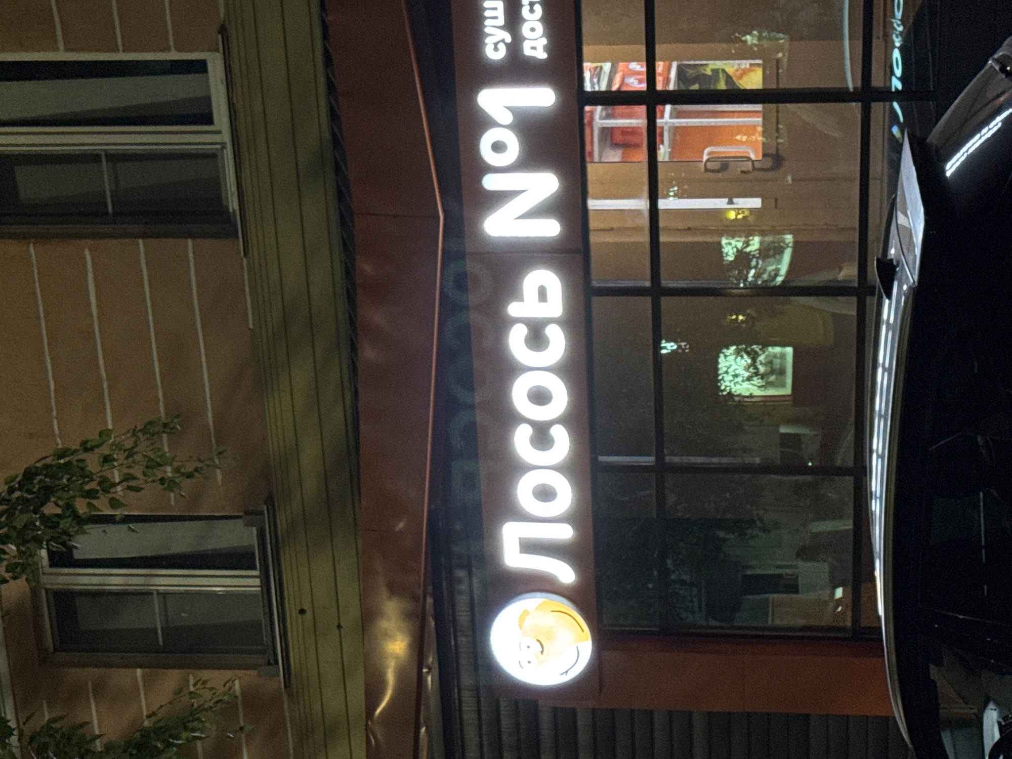 The image shows the facade of a business with a glowing sign that reads "Лосось №1" (Losos No. 1), indicating it is likely a sushi restaurant or a place selling salmon. The sign features a cartoonish smiley face logo to the left of the text. To the right of "№1", there are partially visible words, "суши" (sushi) and "дост" (likely short for доставка, meaning delivery). The sign is illuminated at night, casting a bright white glow. Behind the sign, windows of the establishment are visible, showing some interior lighting and reflections of the exterior. The building has a textured brown facade with horizontal detailing. Below the business, the roof of a car is visible, suggesting it is parked in front of the establishment. The overall setting appears to be a street in the city of Oskemen, Kazakhstan, given the Cyrillic script on the sign. The image is taken at night, with the lighting emphasizing the illuminated sign and the reflections in the windows.