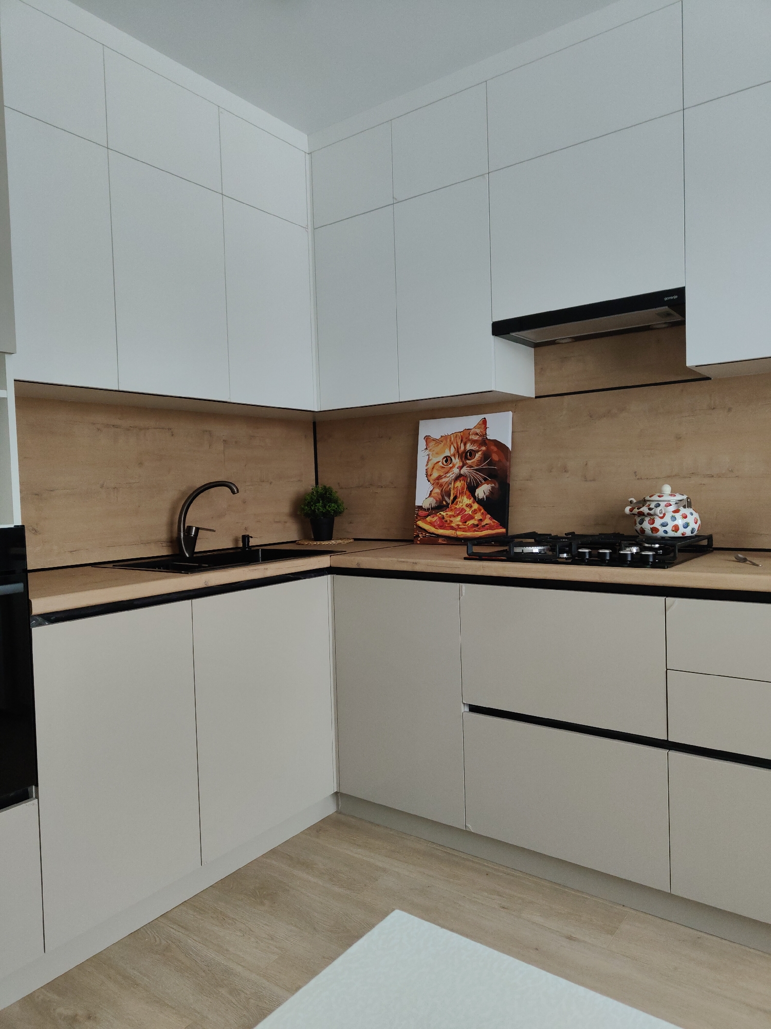 This is an indoor shot of a kitchen with modern design elements. The cabinetry consists of white upper cabinets and light beige lower cabinets, with black hardware accents. The countertops have a wood-grain finish with a black trim. Above the stove is a black range hood. A black sink and faucet are installed on the left counter, with a small potted plant nearby. On the right side, there's a gas cooktop with a colorful teapot to its right, and a framed image of a cat eating pizza is placed behind the cooktop. The flooring is light wood, and a portion of a white surface is visible in the foreground. The location is not specified and is marked as Unknown, Unknown. There are no people visible in the image.