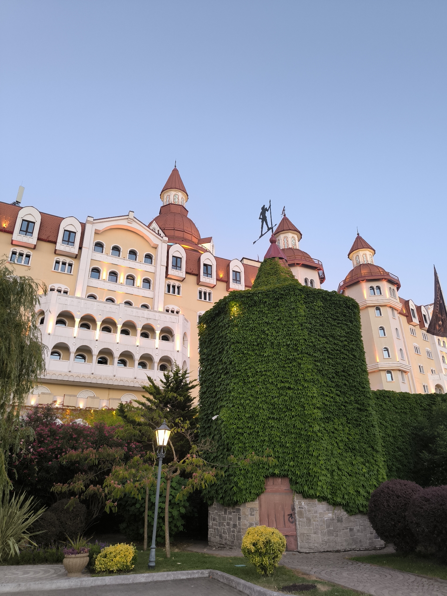 The photograph captures an exterior view of a large, ornate building with multiple towers and red-tiled roofs, characteristic of a castle or grand hotel. The building is primarily yellow with white architectural details, and it is partially covered by lush green ivy. A small, weathered wooden door is visible at the base of the ivy-covered section. In the foreground, a lamppost stands beside a paved walkway and a variety of garden plants, including flowering bushes and manicured shrubs. A silhouette of a figure holding a spear is visible atop one of the towers. The sky is a clear, pale blue, suggesting it is either early morning or late evening. This building is located in Sirius, Russia, and is known as the Grand Hotel Zvezdny.