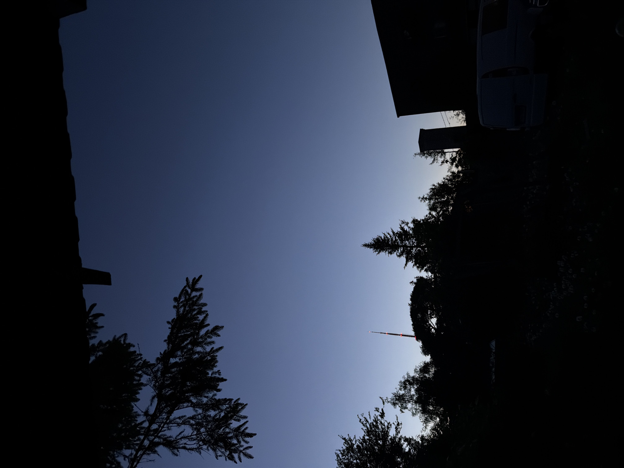 The image depicts an outdoor scene at twilight or pre-dawn, characterized by a clear sky displaying a deep navy blue gradient transitioning to a lighter periwinkle blue at the horizon. The overall lighting renders most foreground and midground elements as dark silhouettes.
In the upper left, a large branch from a coniferous tree, likely a spruce or pine, extends downwards, partially obscuring the sky and an unseen dark structure that appears to be an overhang or roofline.
The horizon is dominated by the silhouettes of numerous trees, a mix of coniferous and deciduous species. Towards the left-center, rising above the tree line, is the distinct silhouette of the Tallinn TV Tower (Teletorn) located in the Pirita district of Tallinn, Estonia. Several red aircraft warning lights are illuminated along the tower's shaft.
To the right, a dark, rectangular building with a distinct chimney structure is silhouetted against the sky. In the lower right corner, a light-colored van, appearing dark grey under the low light, is parked with what appears to be an open or absent side door. The ground level is largely obscured by deep shadow, showing indistinct features that may be vegetation or a garden. No individuals are visible in the scene, and no discernible actions or interactions are taking place.
