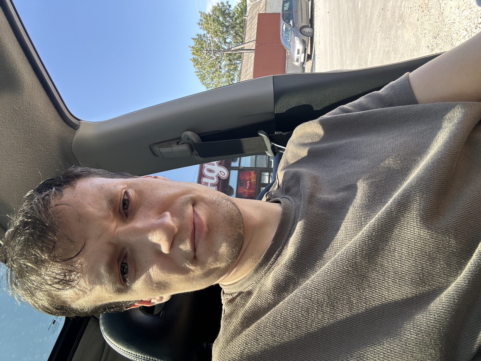 In Shatava, Ukraine, a man is photographed from the chest up, sitting in the driver's seat of a car. The man has short, dark hair and is wearing a brown t-shirt. His gaze is directed towards the camera. The car's interior is visible, with a black headrest on the left and a gray pillar on the right. A seatbelt is fastened to the pillar. Outside the car, through the window, a parking lot can be seen with a gray car parked in the background. Further in the background, a building with a red roof and a red fence is visible, along with trees and a clear blue sky.