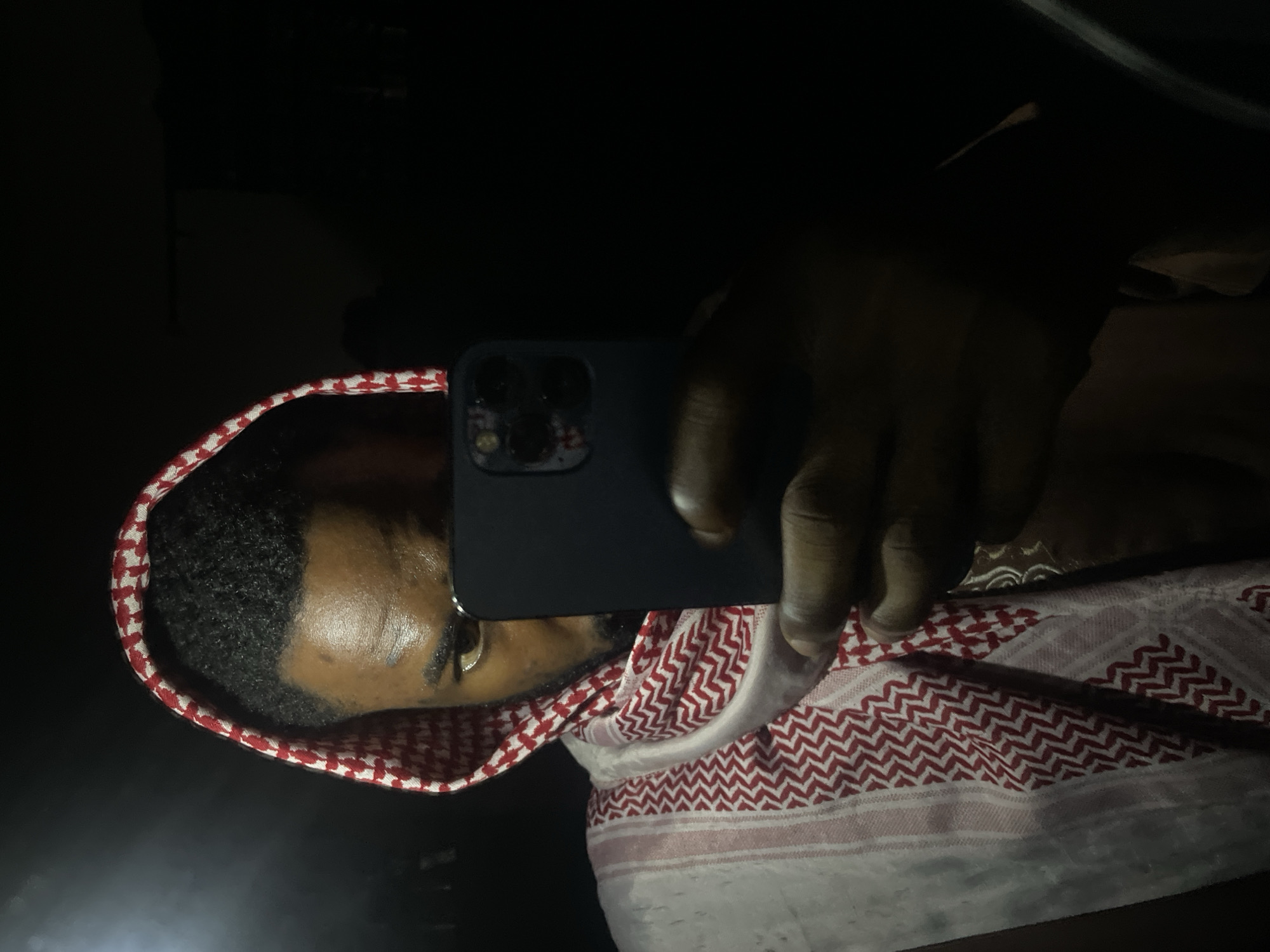 A man wearing a traditional red and white patterned headdress (keffiyeh) is partially visible in the image. He is holding a dark-colored smartphone with three camera lenses up to his face, partially obscuring it. His skin tone appears to be dark. The background is mostly dark and undifferentiated, suggesting low light conditions, possibly indoors or at night. The presence and context of Lokoja, Nigeria, cannot be determined from the image content alone.