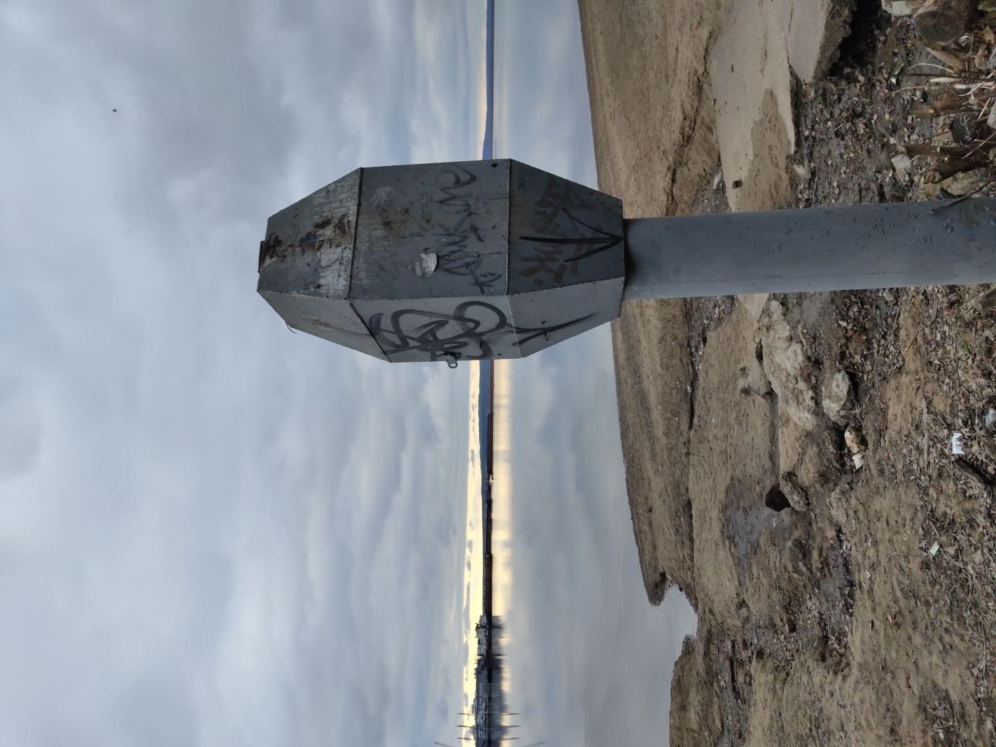 The image displays an outdoor scene, likely along a waterfront in Chaikovsky, Russia. In the foreground, a weathered, grey, octagonal metal object, possibly a utility box or part of a navigational aid, is mounted horizontally on a sturdy grey pole. The metal object is covered with dark, scribbled graffiti.
To the left of the pole and extending into the distance is a calm body of water, which reflects the pale, overcast sky. A distinct, narrow band of bright light stretches across the distant horizon, where the water meets the sky, suggesting either a sunrise or sunset. The sky above is entirely covered with light grey clouds.
To the right of the pole, the ground consists of uneven, light brown earth, appearing sandy or muddy, interspersed with small rocks and debris. The shoreline appears somewhat unmaintained. In the far distance, across the water, very faint outlines of land or distant structures can be discerned. A tiny dark speck, possibly a bird, is visible in the upper left quadrant of the sky. The scene is static, devoid of people or active events, conveying a quiet, possibly somber, mood under an overcast sky.