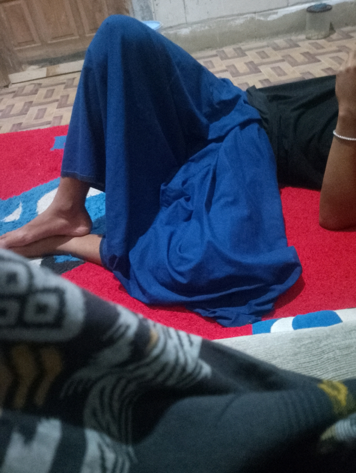 The image depicts a person's legs and feet, partially covered by a blue garment. The person is lying down on a red rug with blue and white patterned sections. The floor in the background appears to be tiled with a geometric pattern, and a wall is visible above it. The scene is indoors, suggesting a domestic setting, potentially in Sowan Kidul, Indonesia, based on the prompt's instruction. A dark garment is visible to the right, and a silver bracelet is on the person's wrist. The foreground is blurred, showing a patterned fabric.