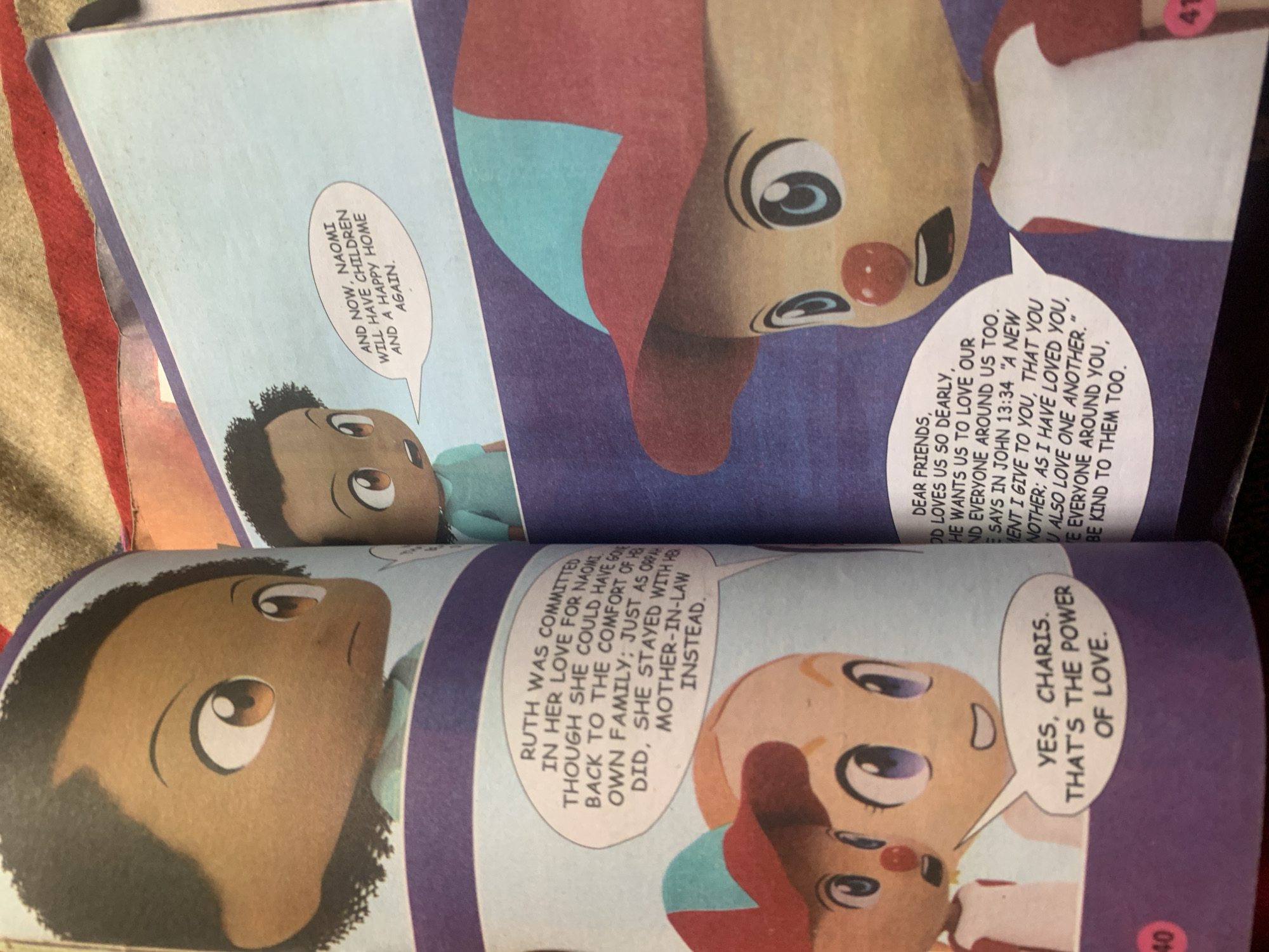 The image displays two pages from a children's book, with cartoon illustrations. On the left page, two young girls with large eyes and simplified features are depicted. The girl on the right has light skin and purple eyes, while the girl on the left has darker skin and brown eyes. They are speaking to each other, with speech bubbles containing text. The girl on the left says, "YES, CHARIS. THAT'S THE POWER OF LOVE."
On the right page, a young boy with dark skin and brown eyes is shown, also with large, cartoonish features. He is speaking, and his speech bubble says, "AND NOW, NAOMI WILL HAVE CHILDREN AND A HAPPY HOME AGAIN." Below him, another cartoon character, a boy wearing a red and teal hat and a white shirt with red sleeves, is speaking, with a speech bubble containing text that appears to be a biblical quote. This character is facing away from the viewer.
The background of the illustrations is colored in muted blues and purples, with some sections featuring a textured appearance suggestive of fabric. The pages are bound together, and a corner of what looks like a woven blanket is visible at the top of the image. A small circle with the number '40' is visible on the bottom left of the left page, and a similar circle with '41' is on the bottom right of the right page. The image does not contain any information about the location in the city of Jauro Yinu, Nigeria.