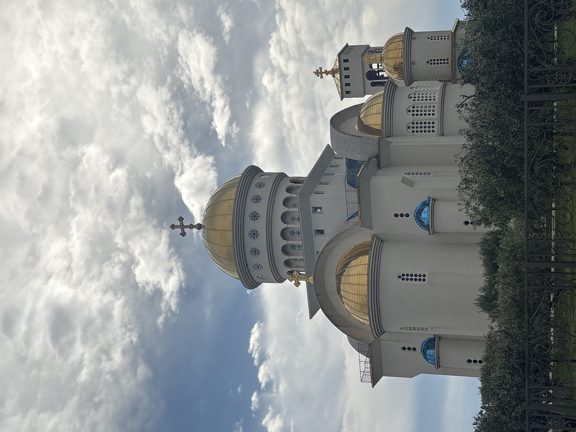 The image showcases the architectural details of a church with golden domes in Bar, Montenegro, taken from a low-angle perspective. The building has a white facade with arched windows and blue accents. The cloudy sky acts as a backdrop. The image is partially obscured by greenery and a black iron fence in the foreground, indicating the structure is situated within a walled or fenced area.