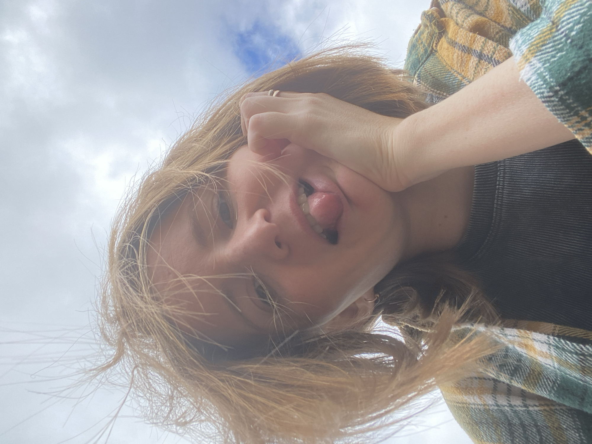 A low-angle, close-up, outdoor selfie depicts a young woman with light brown hair messily styled by the wind. She is wearing a green and yellow plaid shirt over a dark ribbed t-shirt. Her left hand is resting on her cheek, and she is sticking her tongue out at the camera. The background is a cloudy sky with a hint of blue. The image was taken in the city of Chaikovsky, Russia.