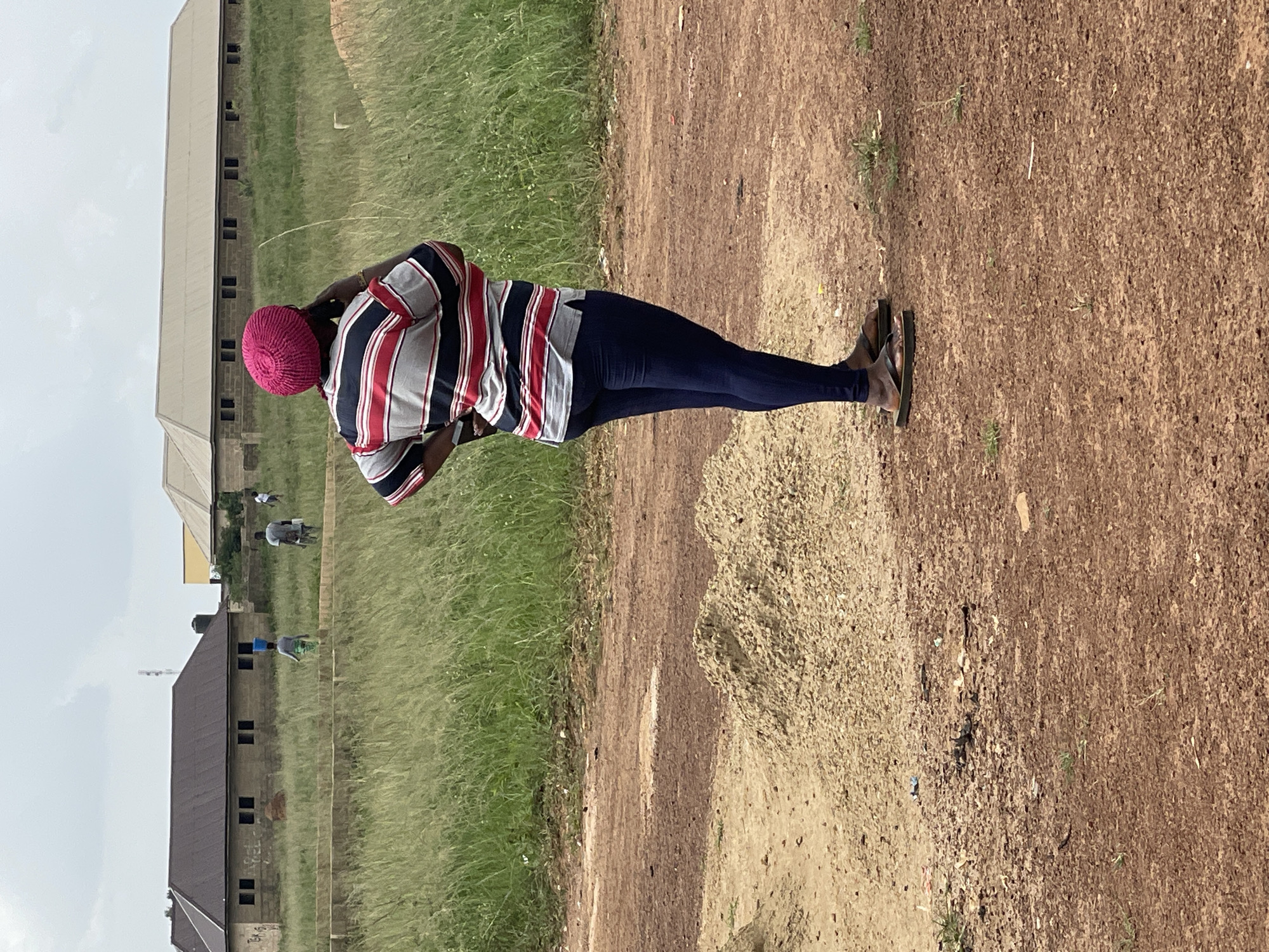 A full shot captures an individual standing in a dirt lot, looking away from the camera and speaking on a mobile phone. The person is wearing a bright pink knitted hat, a horizontally striped t-shirt with red, white, and blue bands, and navy blue leggings. They are also wearing dark brown flip-flops. To the left of the individual, there is a small pile of light-colored gravel. In the background, there are several unfinished buildings with brown roofs and dark windows, set against a backdrop of tall green grass and a cloudy sky. Further in the distance, a cell tower is visible. The scene appears to be an undeveloped area in Durumi, Nigeria, possibly a construction site, with evidence of building materials and uneven terrain.
