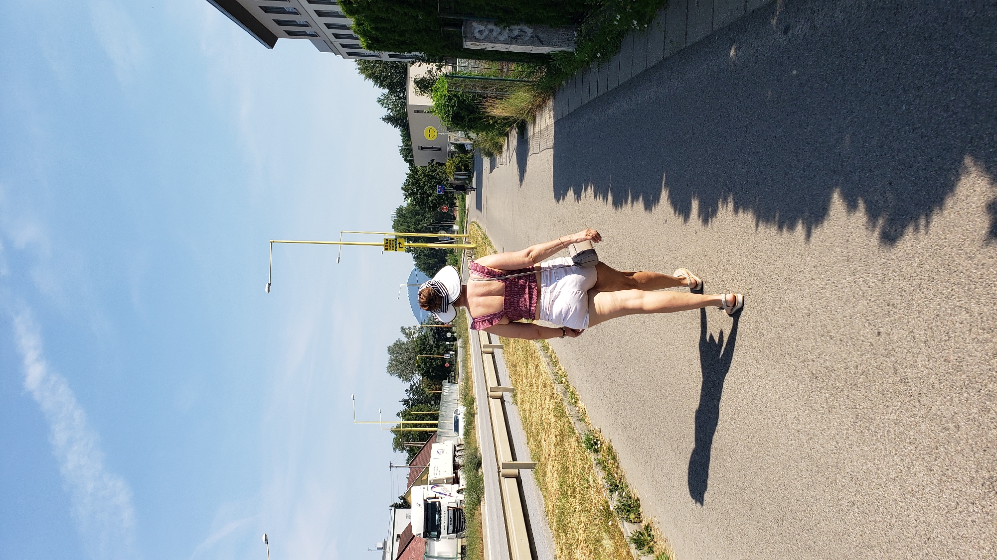 A woman with dark hair pulled up is walking away from the camera on a paved sidewalk. She is wearing a pink ruffled crop top, white shorts, a wide-brimmed white sun hat, and tan sandals. She is also carrying a small gray purse on her right shoulder. The woman is walking on a paved path that runs alongside a road. To her left, there is a divided highway with a guardrail and dry grass. A white truck is visible on the highway. To her right, there is a grassy area with trees and a modern building with a dark roof. The sky is clear and blue with a few wispy clouds. The scene appears to be a suburban or semi-industrial area on a sunny day. The specific location is near the entrance to the industrial park in Prešov, Slovakia, indicated by the yellow street signs with Slovakian text.