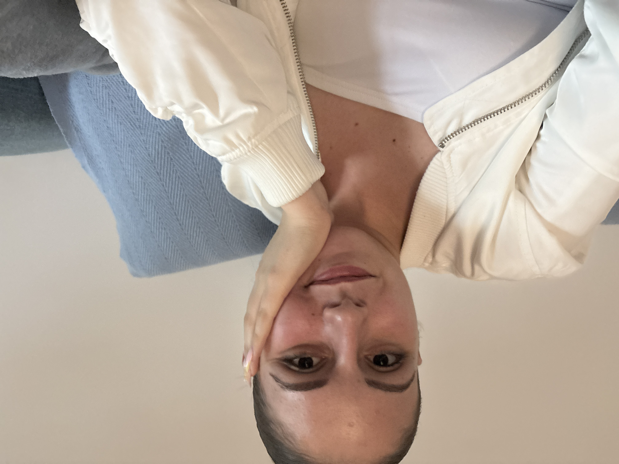 A woman is seen in an upside-down close-up shot. She is fair-skinned with dark eyes and hair pulled back. She is wearing a white top and a white bomber jacket that is partially unzipped. Her left hand is resting on her cheek. The background includes a light blue and gray blanket and a neutral-toned wall. There is no indication of Burgas, Bulgaria in the image.