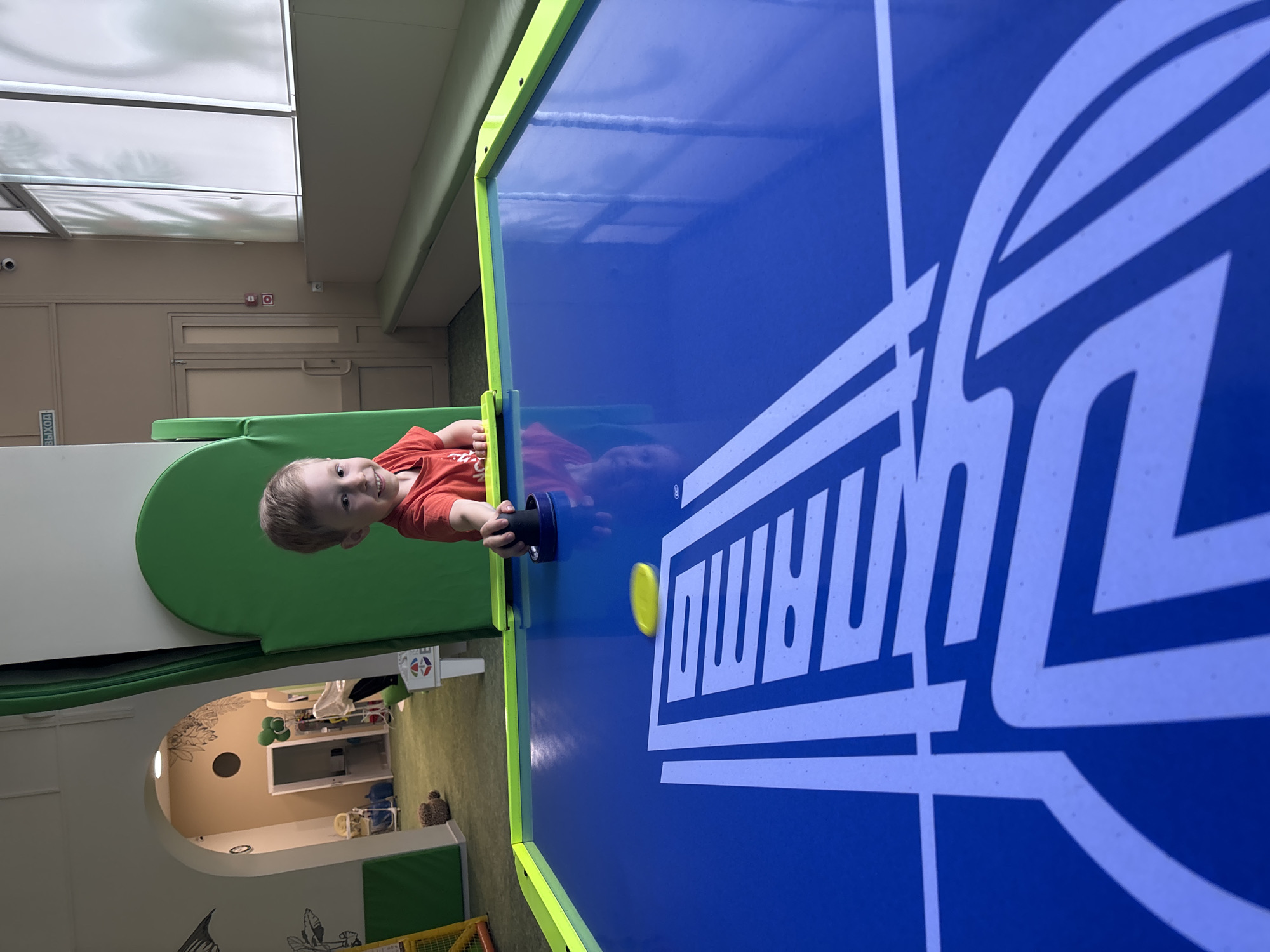 A young boy plays air hockey in an indoor play area. He wears a red shirt and holds a black air hockey paddle in his right hand. A yellow air hockey puck is in play on the blue air hockey table. The location appears to be an indoor play area in a commercial space. In the background is what looks to be a daycare area with toys and a decorated wall. Based on the tags, this is in the city of Chaikovsky, Russia.