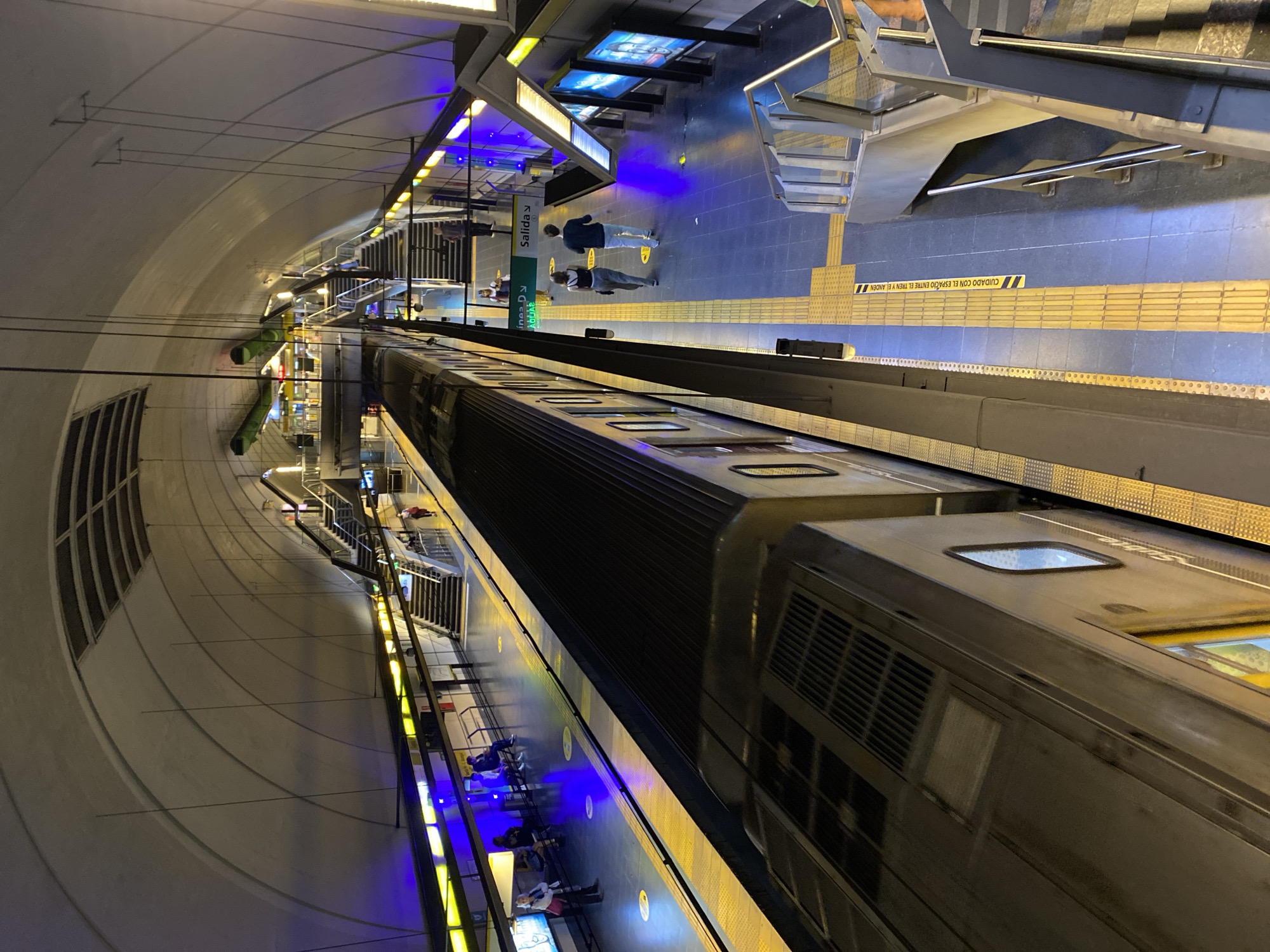 The image depicts a subway station in Buenos Aires, Argentina. The station has a curved ceiling and is brightly lit with a combination of white and blue fluorescent lights. A long subway train is visible along the platform. There are people waiting for the train or walking around the platform area. The platform has tactile paving to aid visually impaired individuals. There are signs in Spanish. Stairs and escalators provide access to other levels of the station.