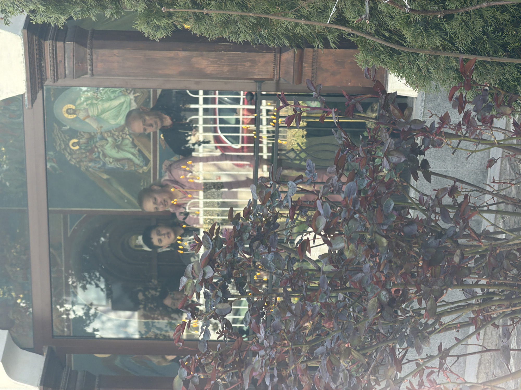 The image shows a building with a large window reflecting people and religious iconography. The window frame is dark wood. Behind the window, there are metal bars. The people in the reflection appear to be inside the building. The iconography includes a depiction of Jesus Christ. In the foreground, there is a bush with red and purple leaves. Above the building, there is a coniferous tree. The building is possibly located in Sofia, Bulgaria, based on the architecture and style of iconography.