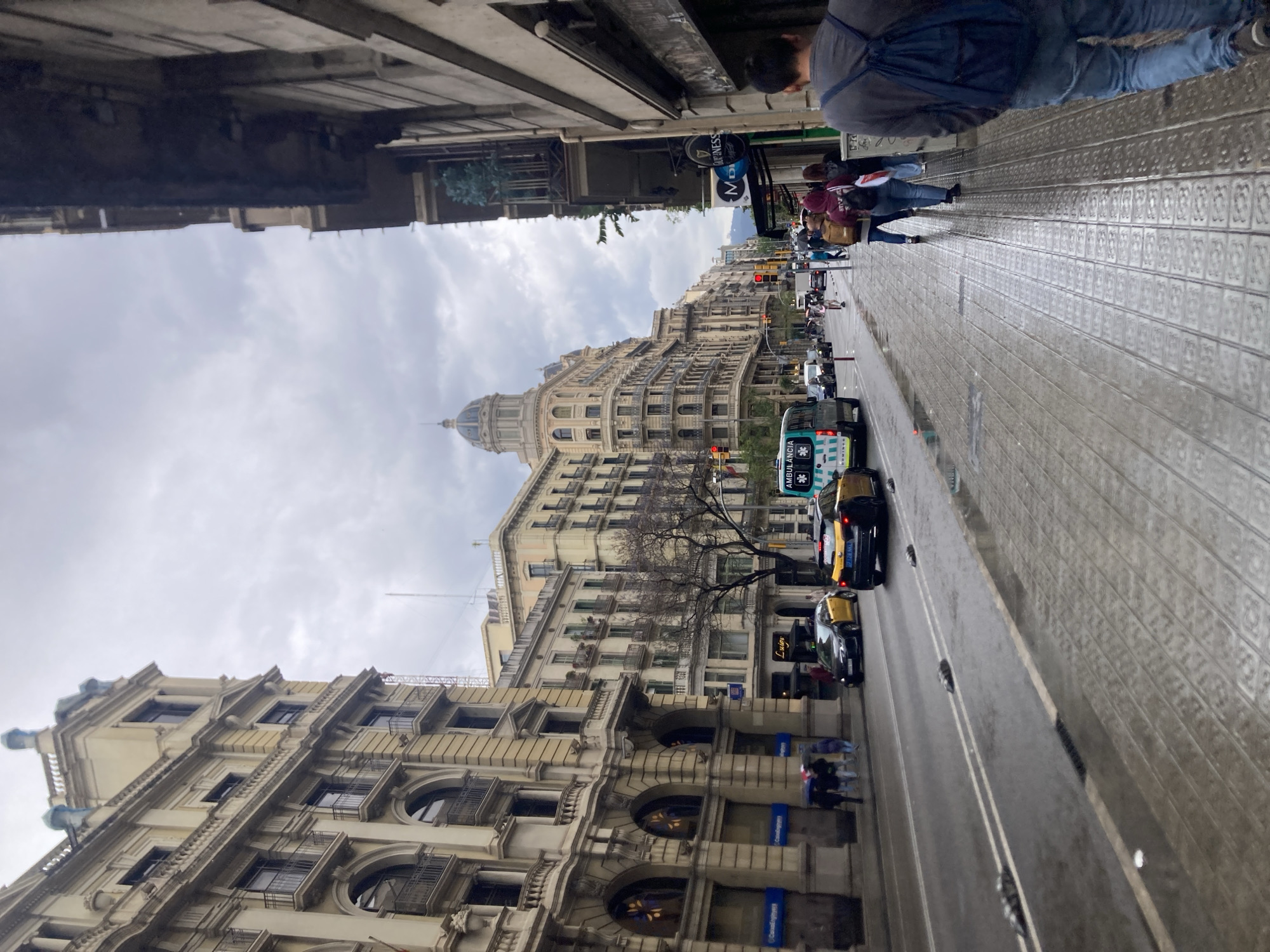The image shows a street scene in Barcelona, Spain. The view is from a sidewalk lined with patterned tiles, looking down a wide avenue flanked by tall, ornate buildings. The buildings are primarily beige and feature multiple stories with numerous windows, balconies, and architectural details like arched windows and decorative cornices. A large domed building is visible further down the street.
The sky is overcast, suggesting it may be a cloudy or rainy day. The street is damp, reflecting light. Several cars and an ambulance are traveling down the avenue. Pedestrians are walking along the sidewalk. There's signage indicating businesses along the street. The overall scene captures the urban environment of a European city street with a mix of vehicular and pedestrian traffic.
