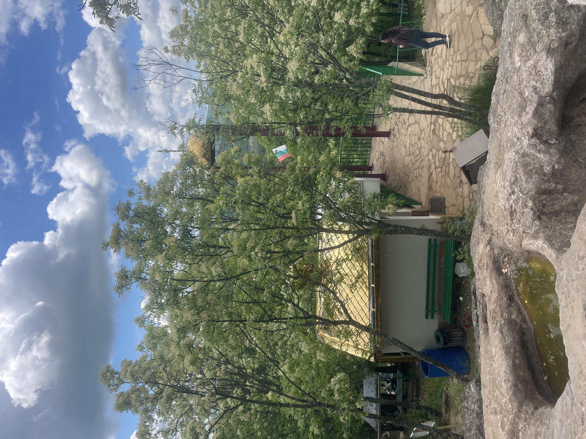 The image depicts a daytime outdoor scene in Novo Selo, Bulgaria, featuring a variety of elements. On the right is a large rock formation with a basin containing water. A building is visible in the mid-ground with a gold-colored roof and white walls, partially obscured by trees. A green bench is situated near the building, along with a blue container. A flagpole with a Bulgarian flag is present near the top of the frame. A woman is partially visible walking across a stone-paved area. The sky above is blue with white clouds.